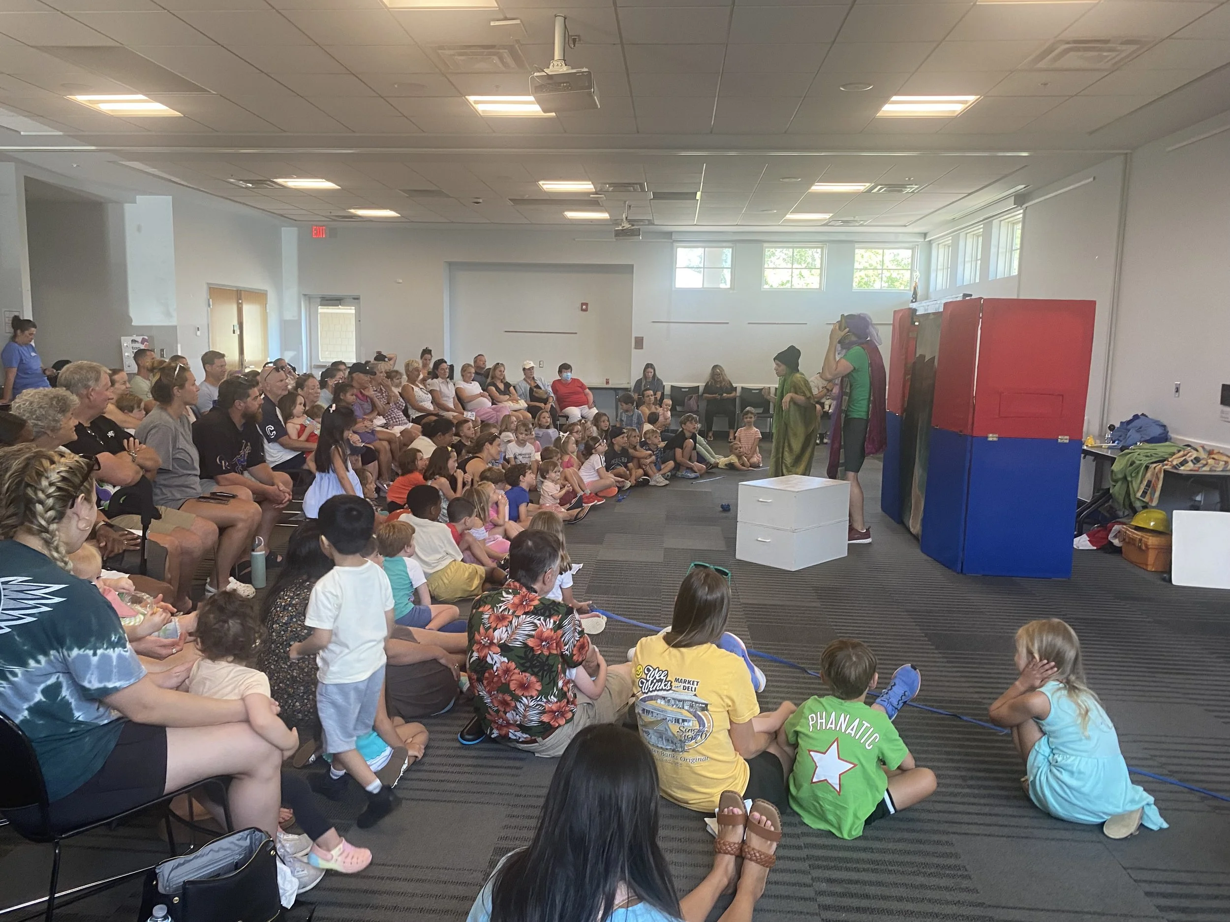 A large group of children and adults watching a clown and a performer in colorful costumes in a room with a gray carpet, white walls, and large windows.