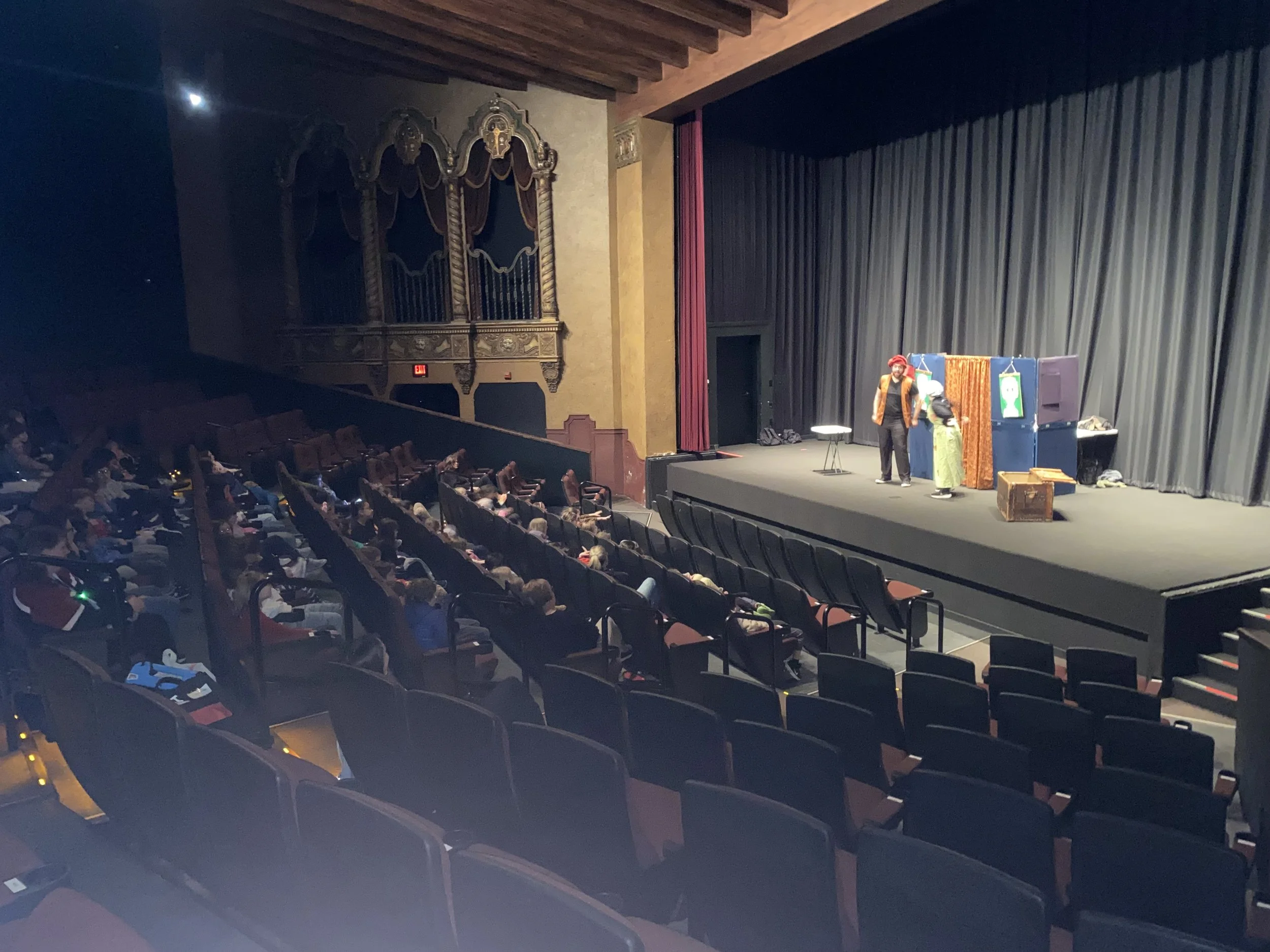 Theater stage with two actors performing in front of an audience seated in a darkened auditorium. The stage has a black curtain background and minimal props, including a small table, a crate, and some framed artwork.