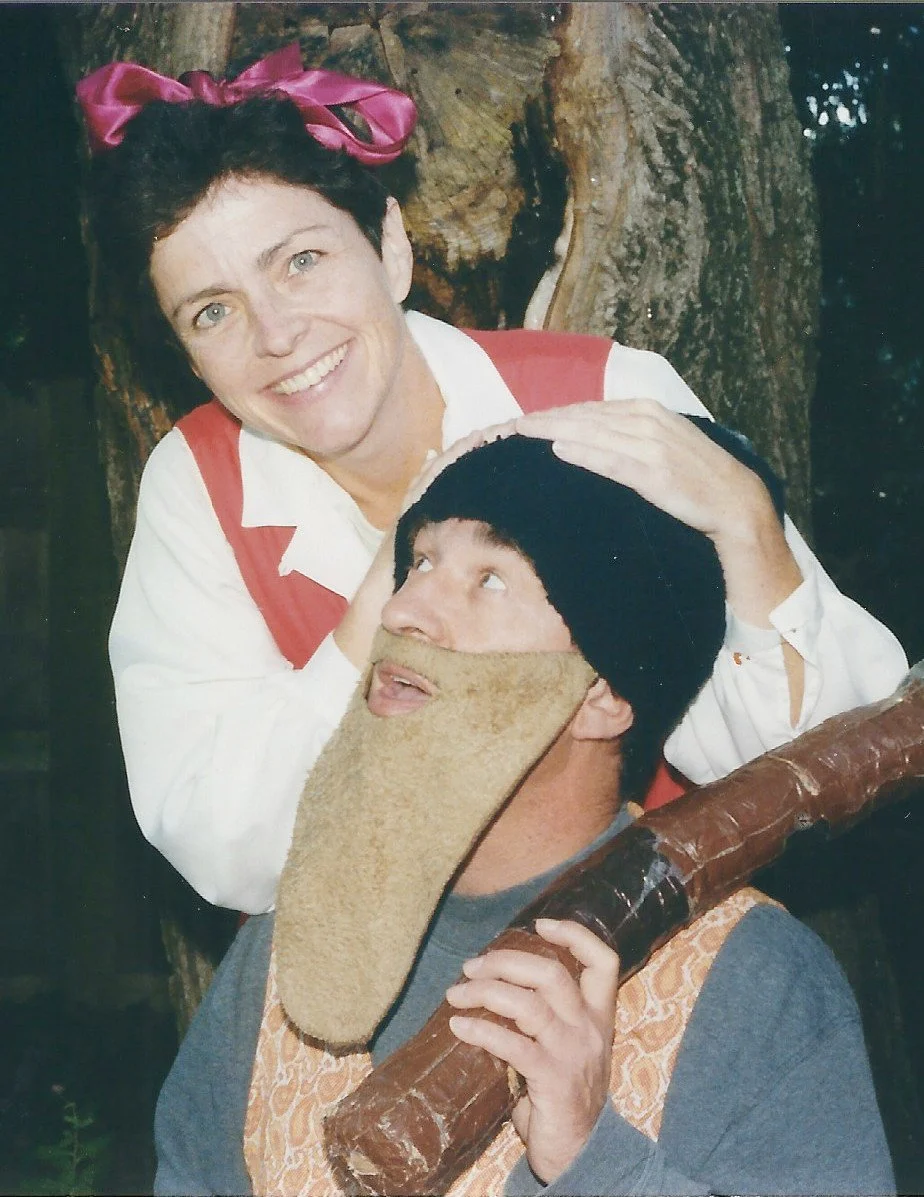 A woman with short dark hair, wearing a pink bow, and a white and red shirt, smiling, has her hands on a man's head. The man has a beard, a black beanie, a gray shirt, and a strange beard made of a cloth or material. He is holding a large stick or staff. They are in front of a tree, outdoors.