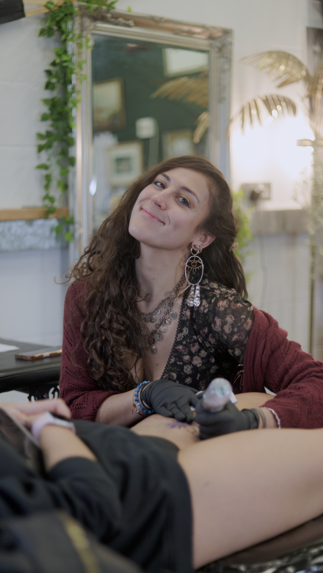 A young woman with long curly hair, jewelry, and earrings is smiling while tattooing someone's leg in a tattoo studio with plants and a decorative mirror in the background.