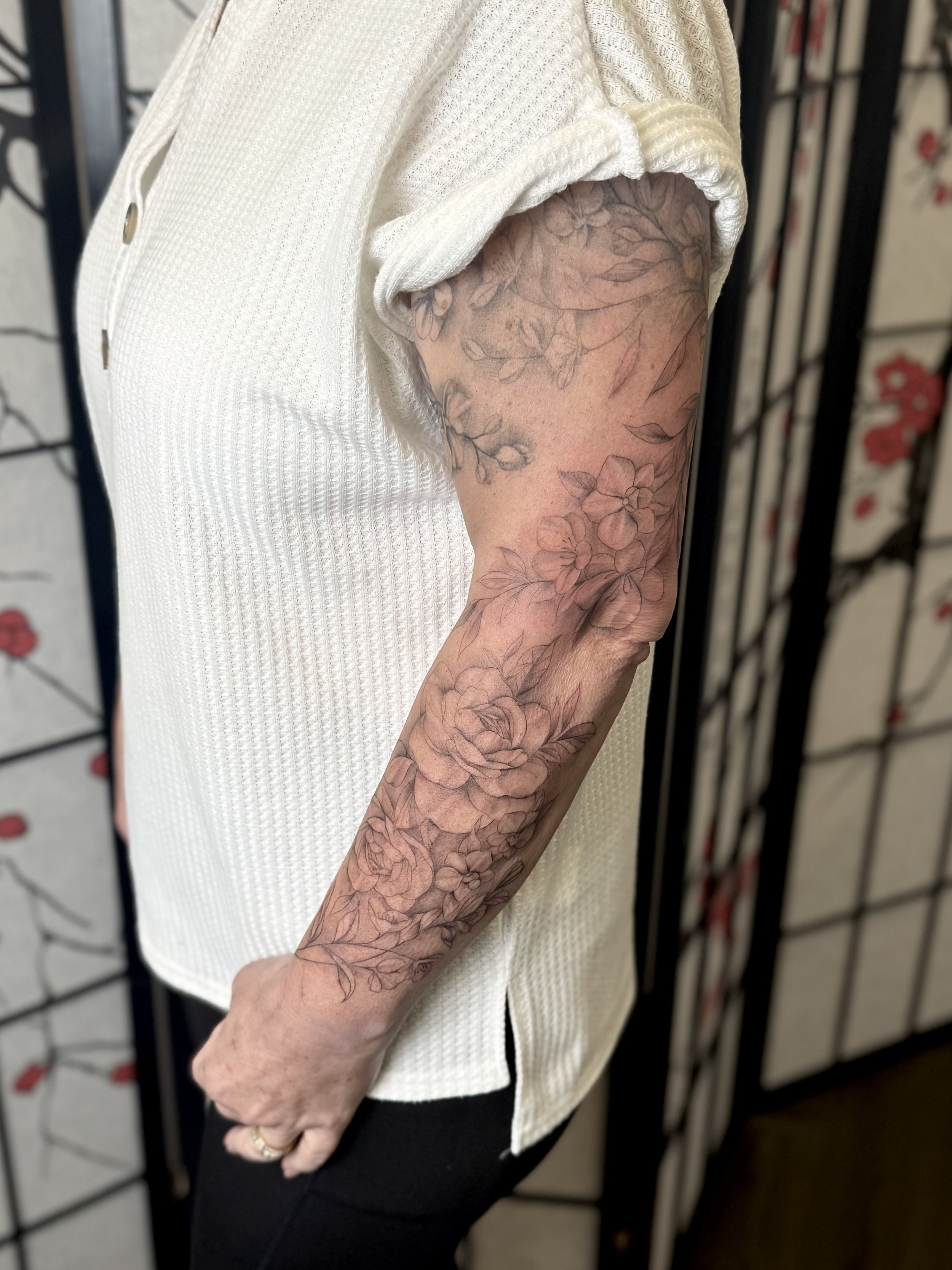 A person's arm with floral tattoos, wearing a white short-sleeve shirt, standing in front of a decorative folding screen.