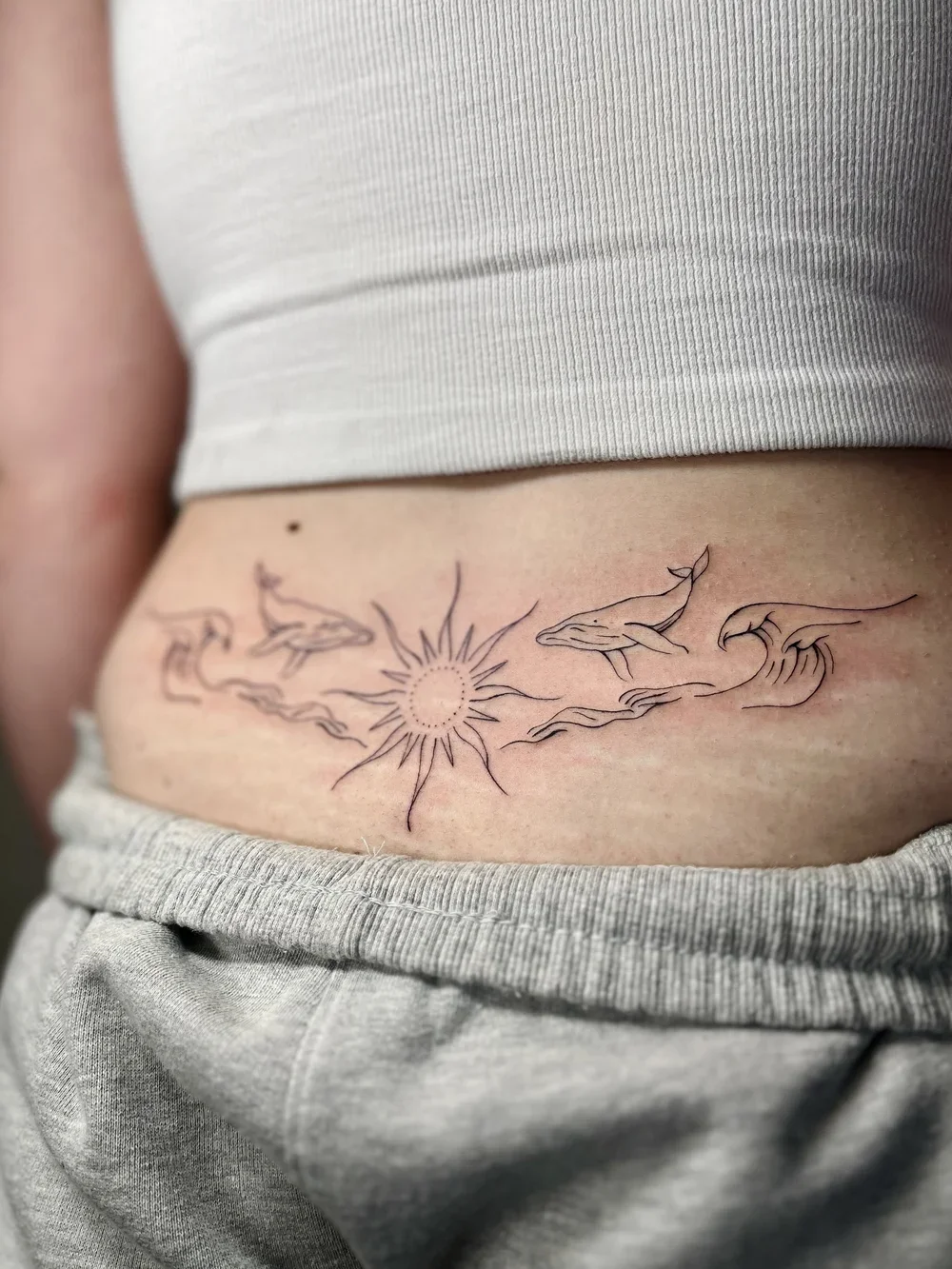 A tattoo of a sun with rays, surrounded by two whales and a wave, located on a person's lower abdomen.