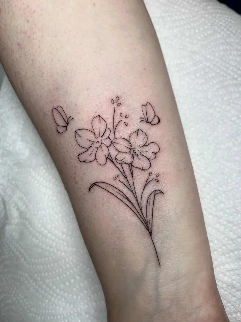A tattoo of a simple black outline of a daffodil flower with two butterflies surrounding it on a person's forearm.