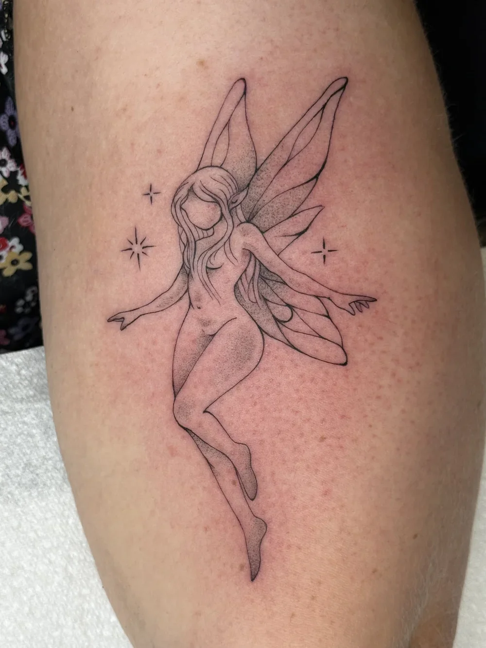 Line art tattoo of a fairy with long hair and butterfly wings, standing with arms outstretched, surrounded by small star-like sparkles on skin.