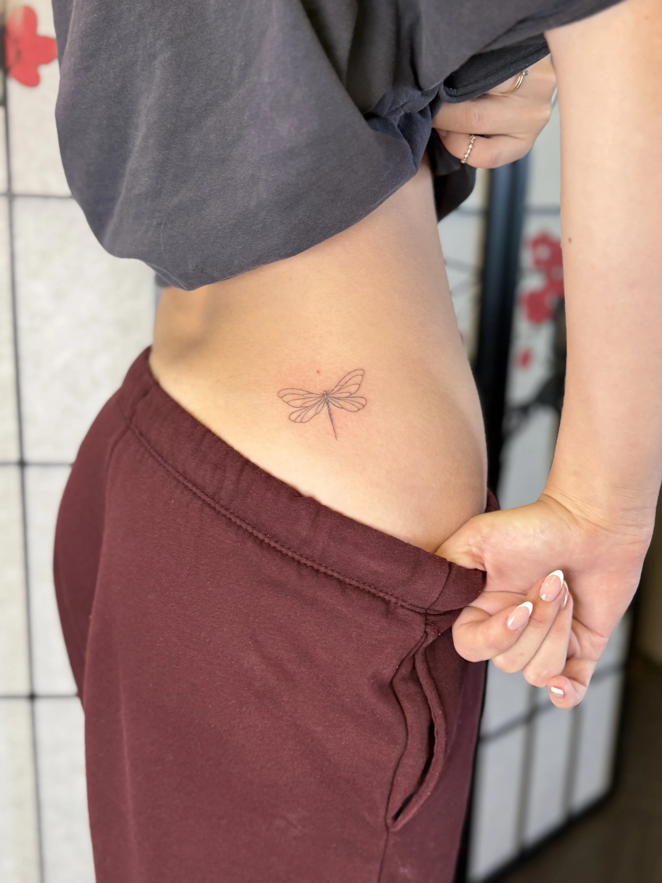 Person pulling down maroon pants to reveal a small dragonfly tattoo on their side near the waist, with a gray shirt and a background with a floral pattern.