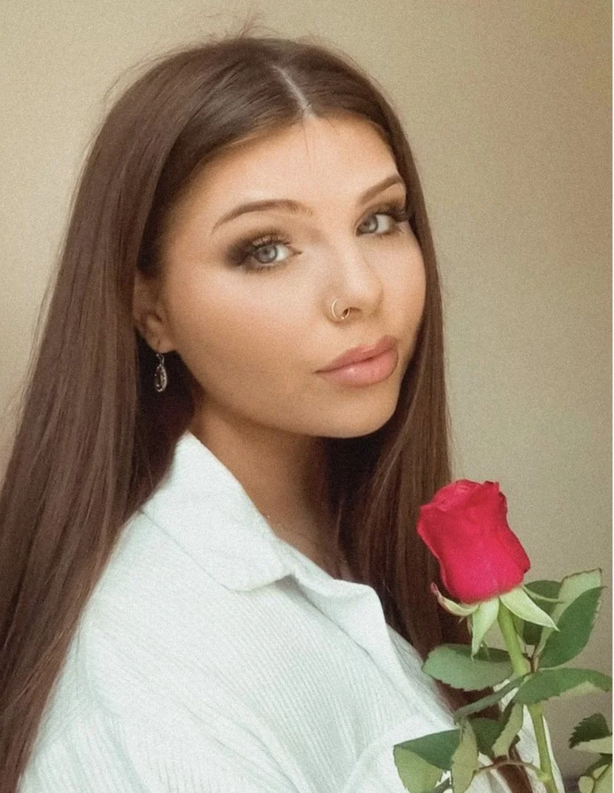 A woman with long brown hair and blue eyes holding a red rose, wearing a white shirt and silver hoop earrings, standing against a beige background.