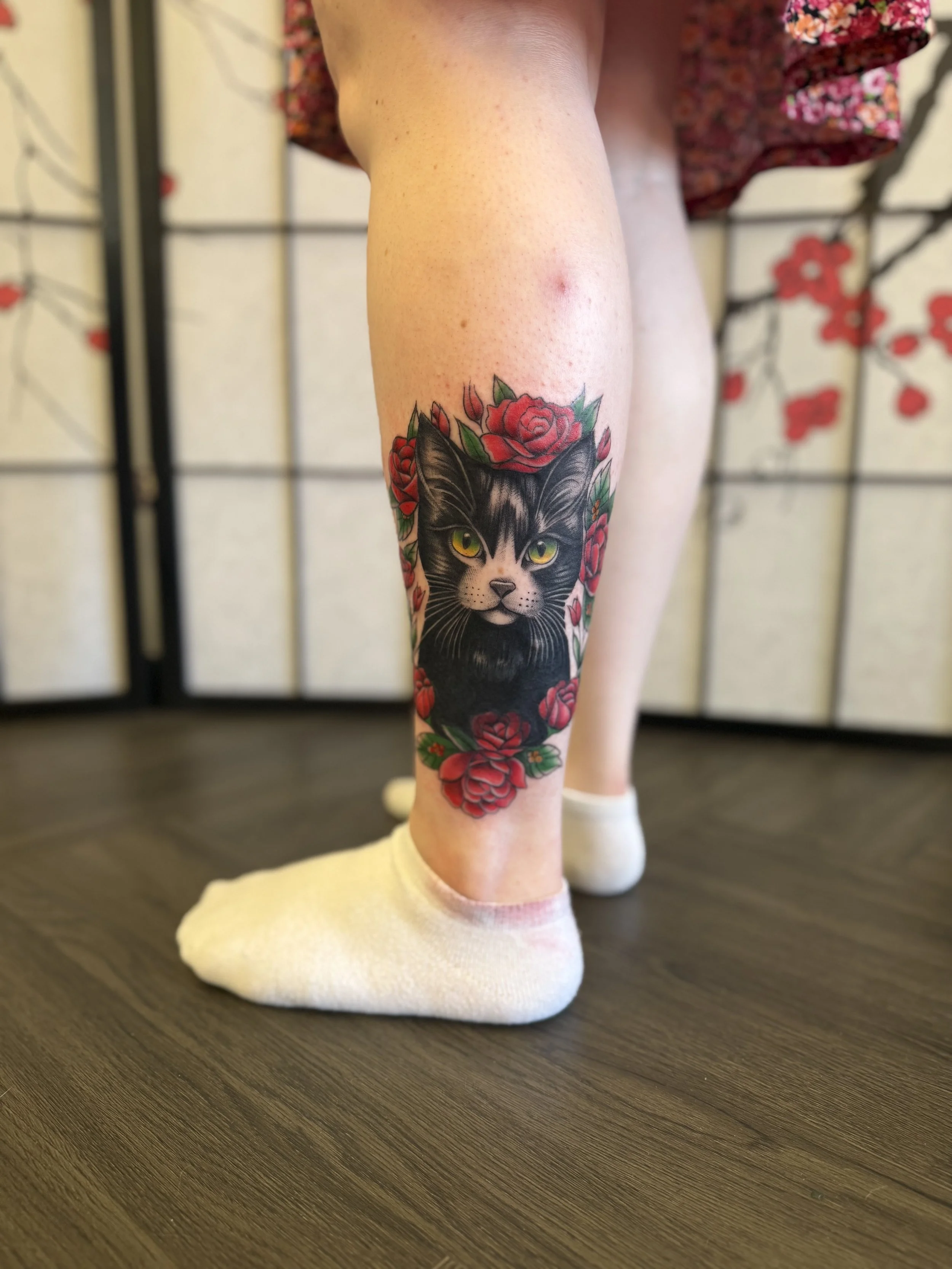 Tattoo of a black and white cat with yellow eyes, surrounded by red roses, on a person's lower leg.