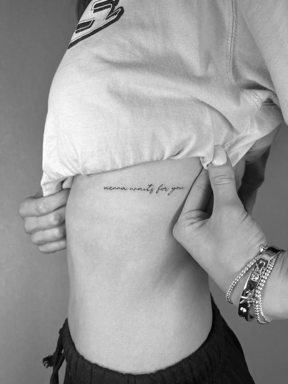 A person's side waist with a tattoo reading "vienna waits for you". The person is wearing a loose t-shirt and dark shorts, with a hand resting on their waist. The person is wearing bracelets and has a manicured thumbnail.