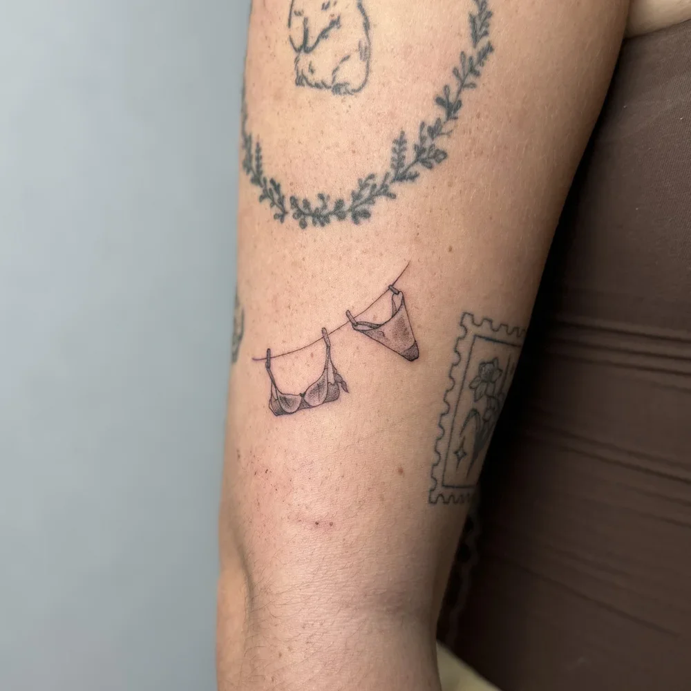 Tattoo of a clothesline with a bra and a pair of underwear hanging on it on a person's arm.
