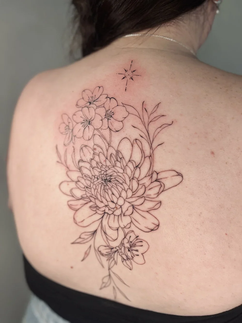 A person's shoulder with a fresh, black ink outline floral tattoo featuring various flowers and leaves, and a small star or compass-like design near the neck.