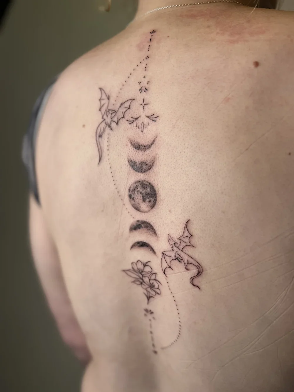 A tattoo on a person's shoulder featuring lunar phases, planets, a floral element, bats, and dotted lines with decorative details.