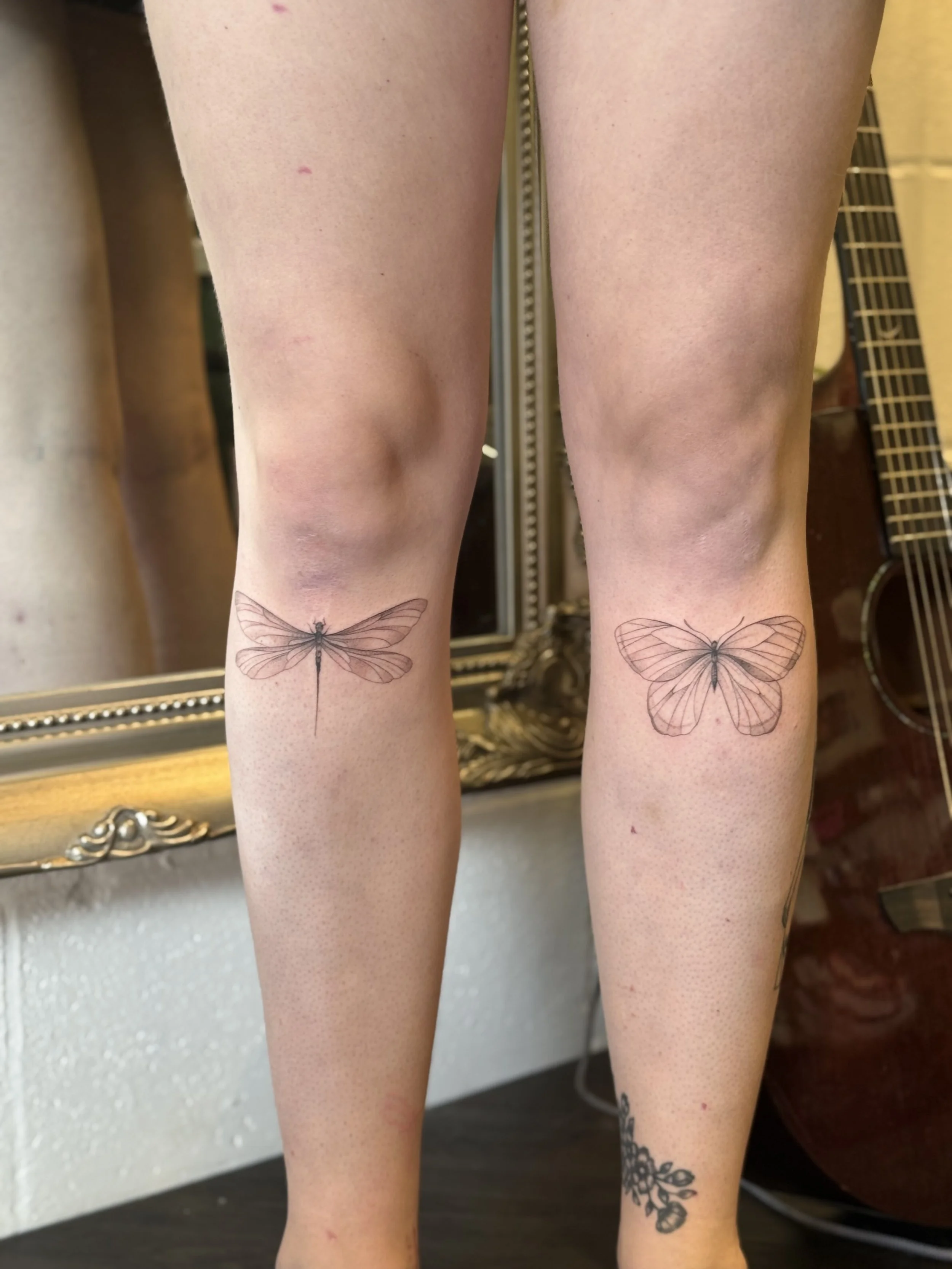 Tattooed knees with butterfly designs on the front of the legs, with a mirror and guitar in the background.