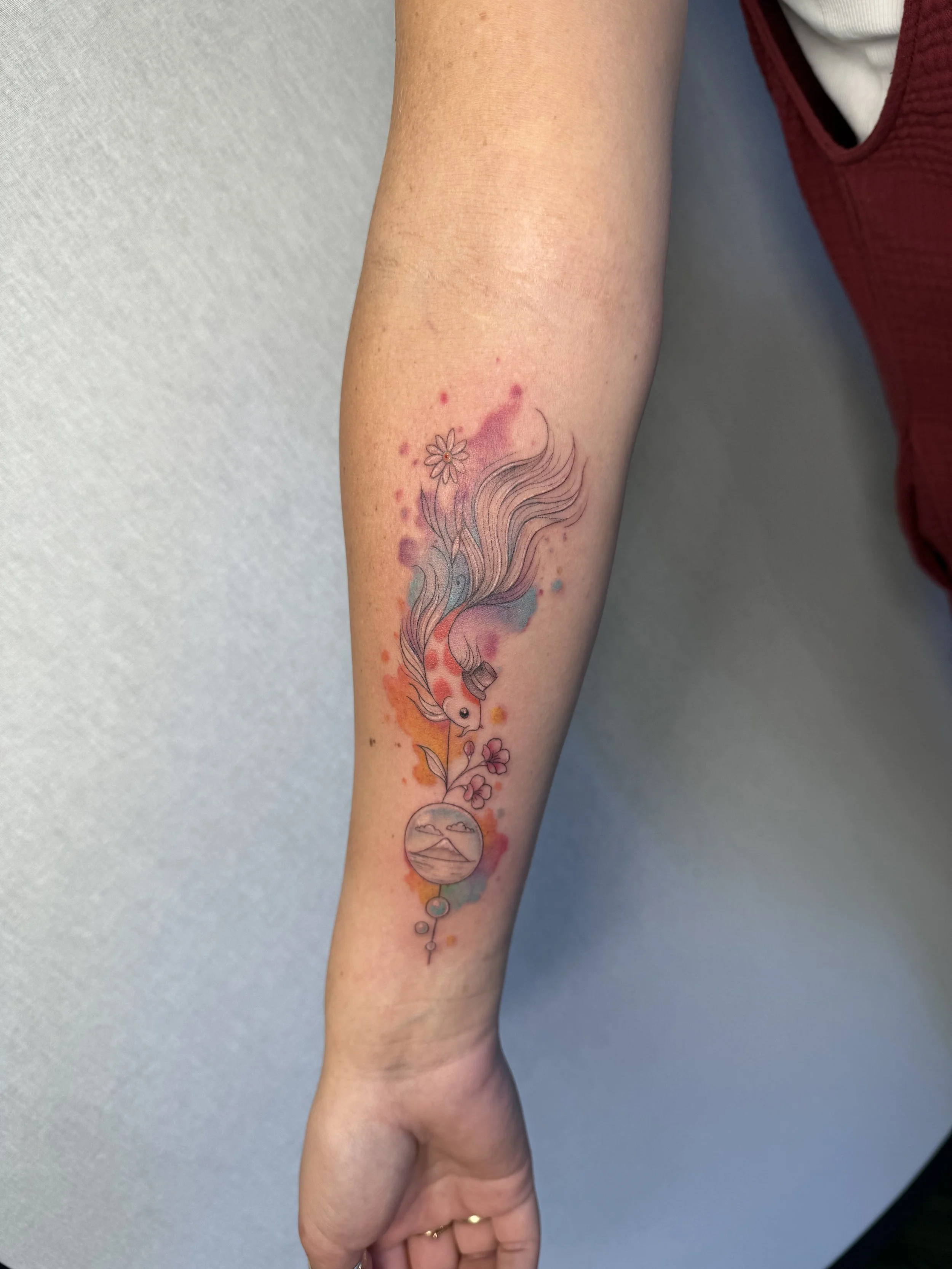 Tattoo of a colorful fish with flowing fins, surrounded by flowers and a circular sunset landscape, on a person's forearm.