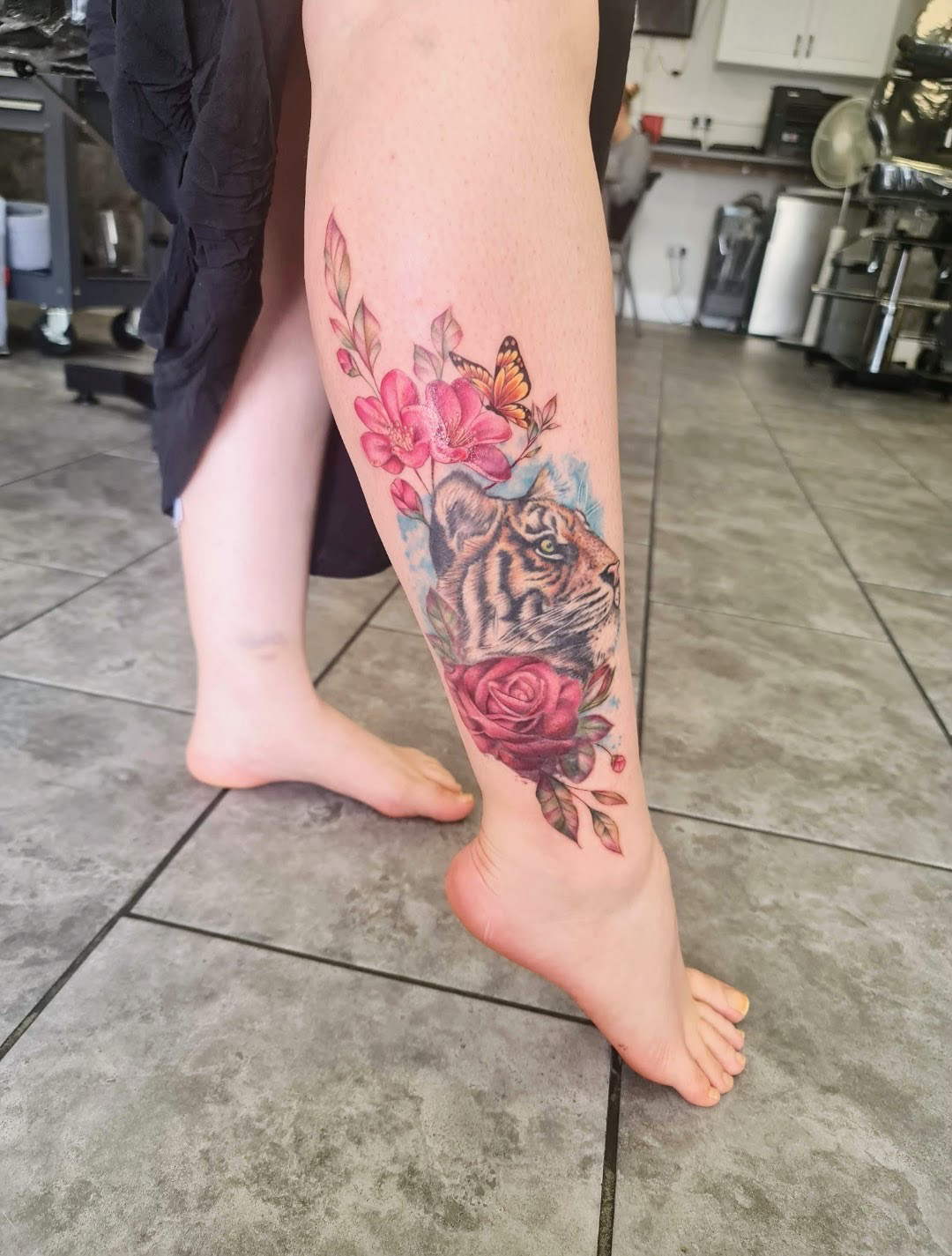 A person with a tattoo of a tiger's face surrounded by pink flowers and a butterfly on their thigh and calf.