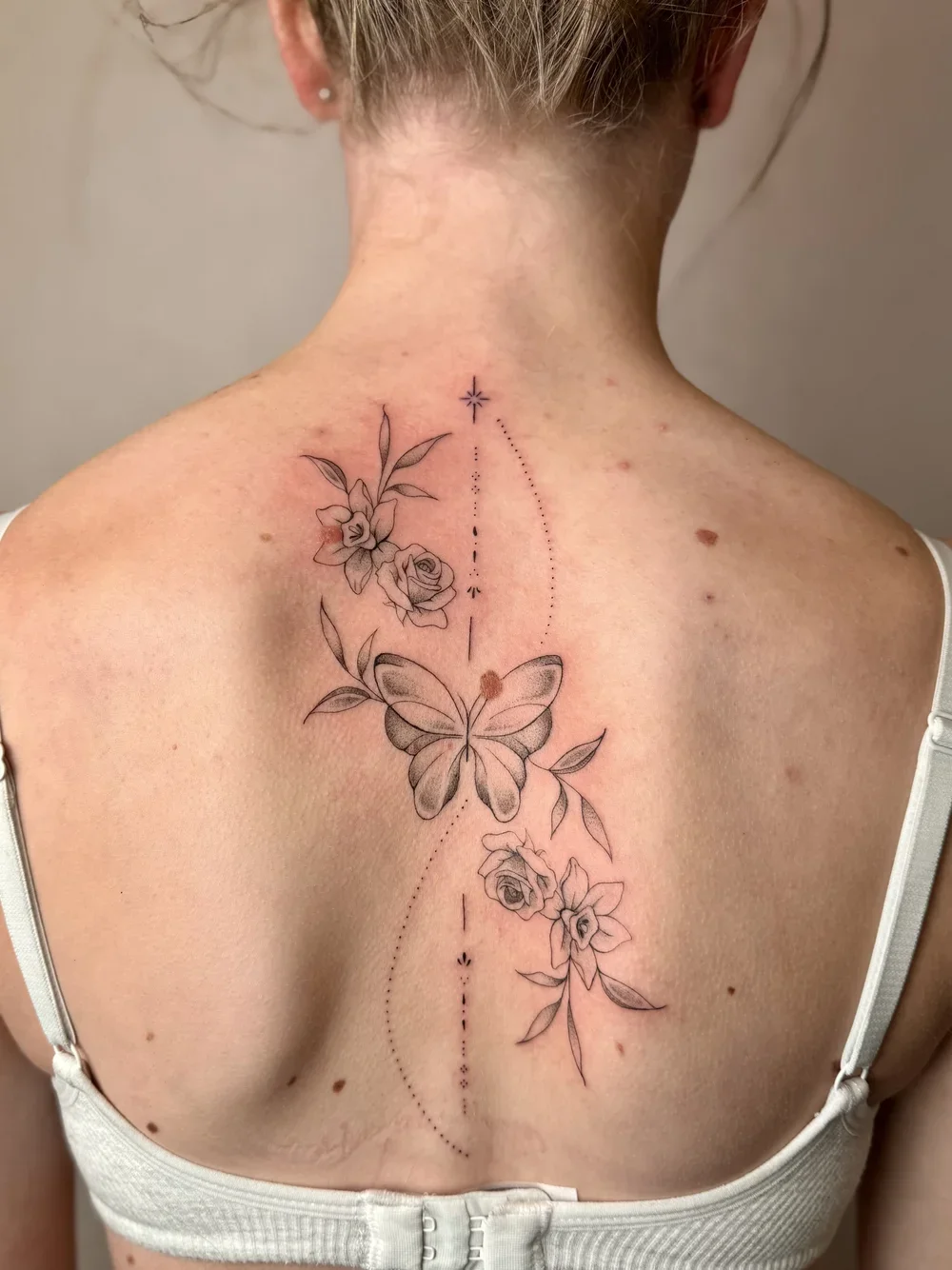A tattoo on a person's back featuring a butterfly surrounded by flowers and decorative lines, with dotted and solid lines and small symbols.