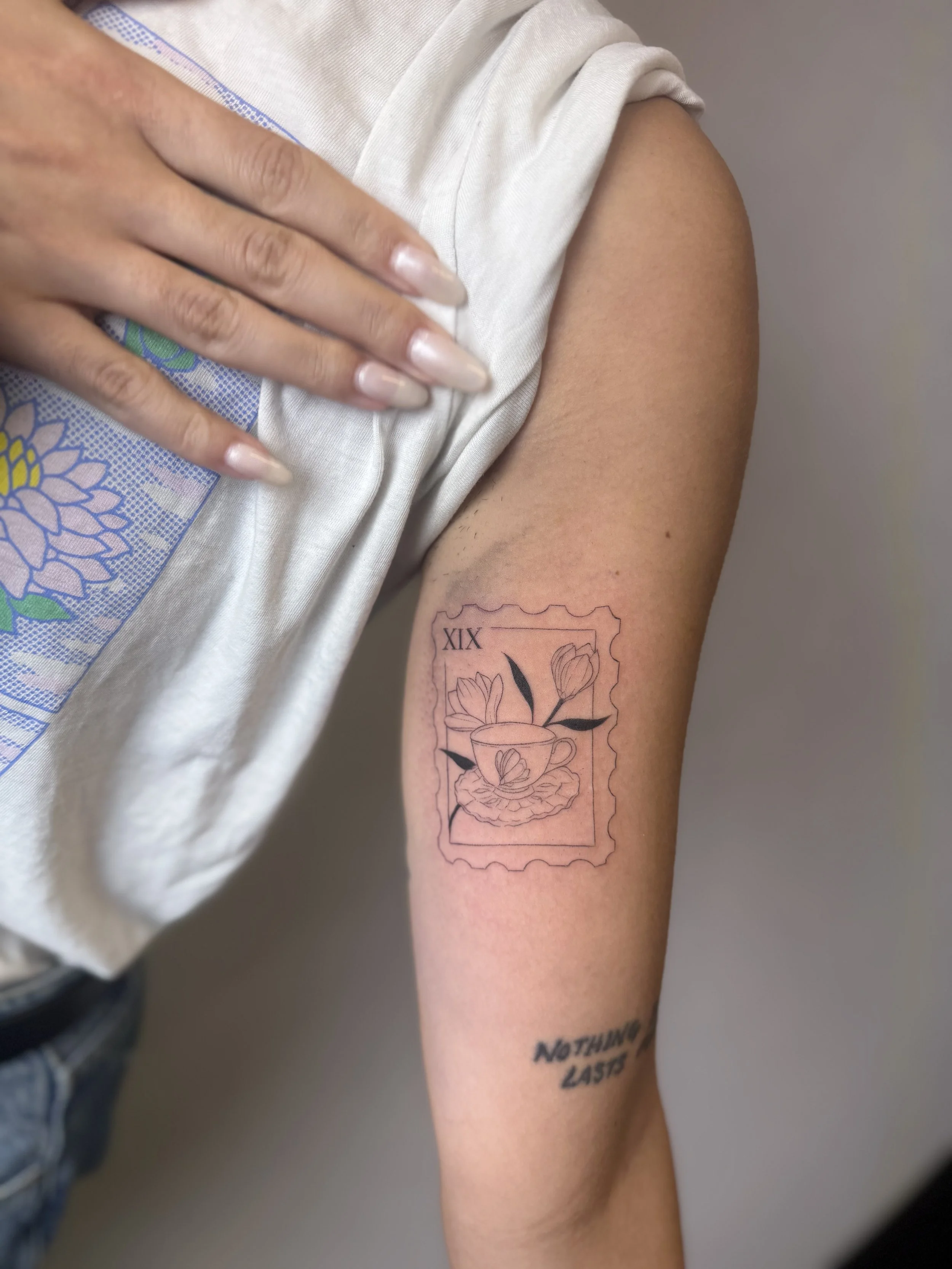 Tattoo of two tulips and a butterfly inside a postage stamp outline on a person's arm, with the Roman numeral XIX and the phrase 'Nothing Lasts' below.