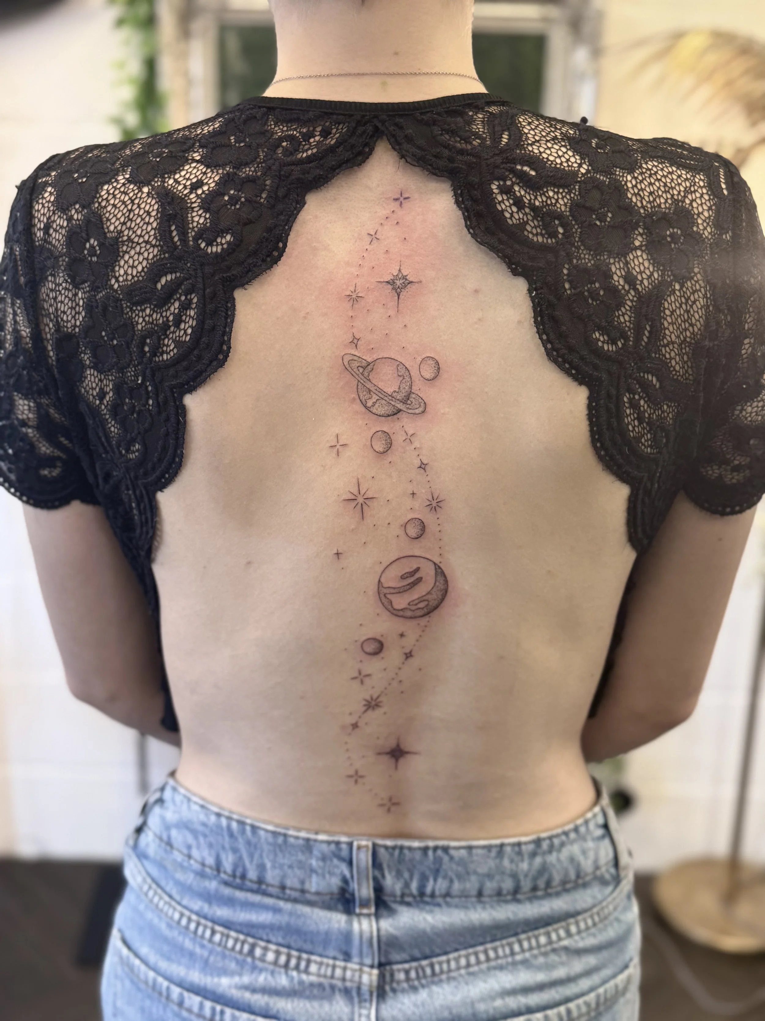 A person with a space-themed tattoo running vertically down their back. The tattoo features planets, stars, and celestial designs in a delicate, minimalist style.