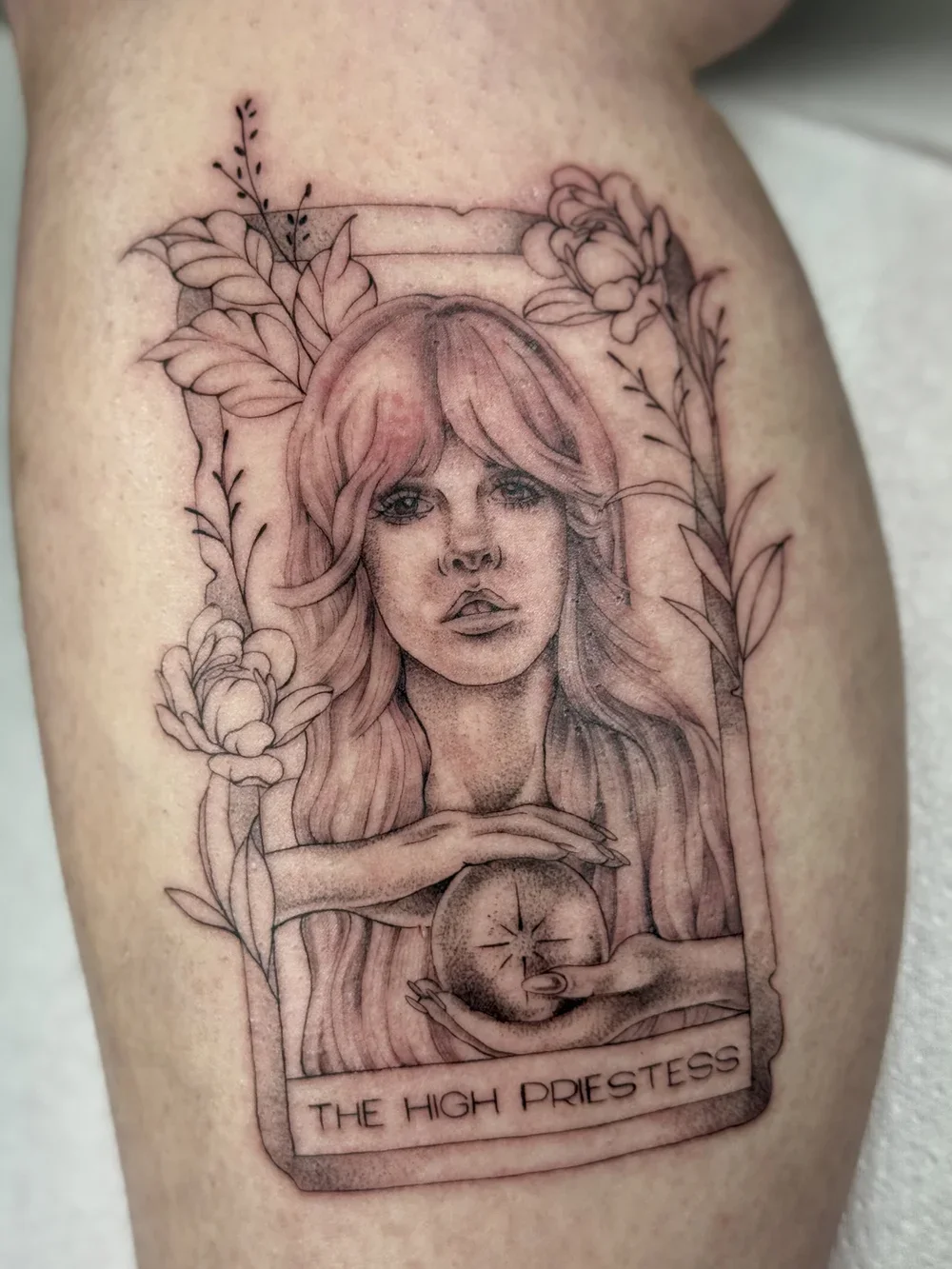 Tattoo of a woman with long hair surrounded by flowers, holding a crystal ball, with the caption 'The High Priestess' at the bottom.