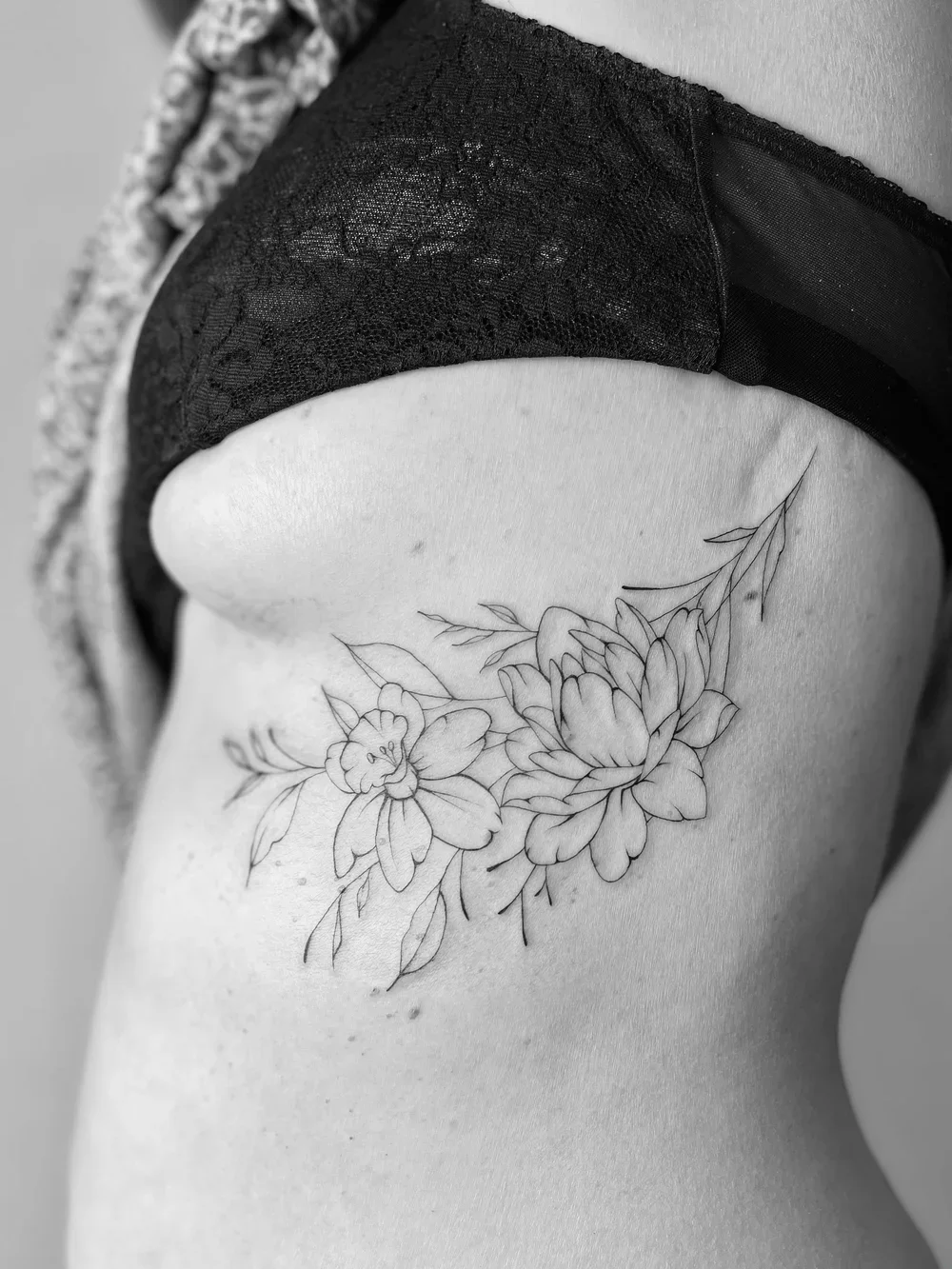 Black lace underwear worn by a person with a floral tattoo on their side, featuring detailed flowers with leaves.