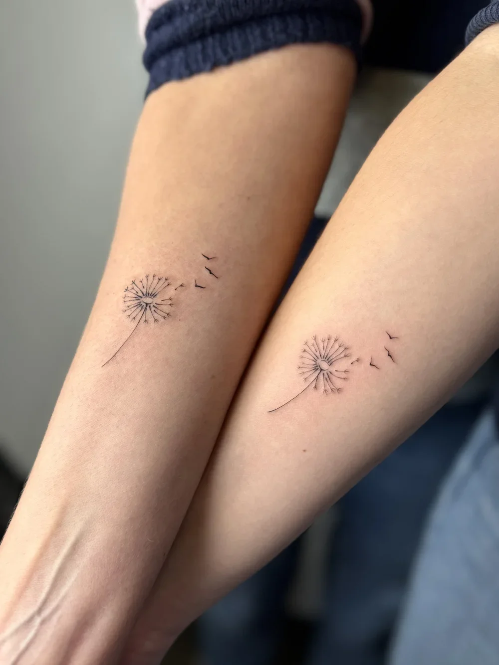 Two forearms with matching dandelion tattoos that appear to be dispersing into birds.