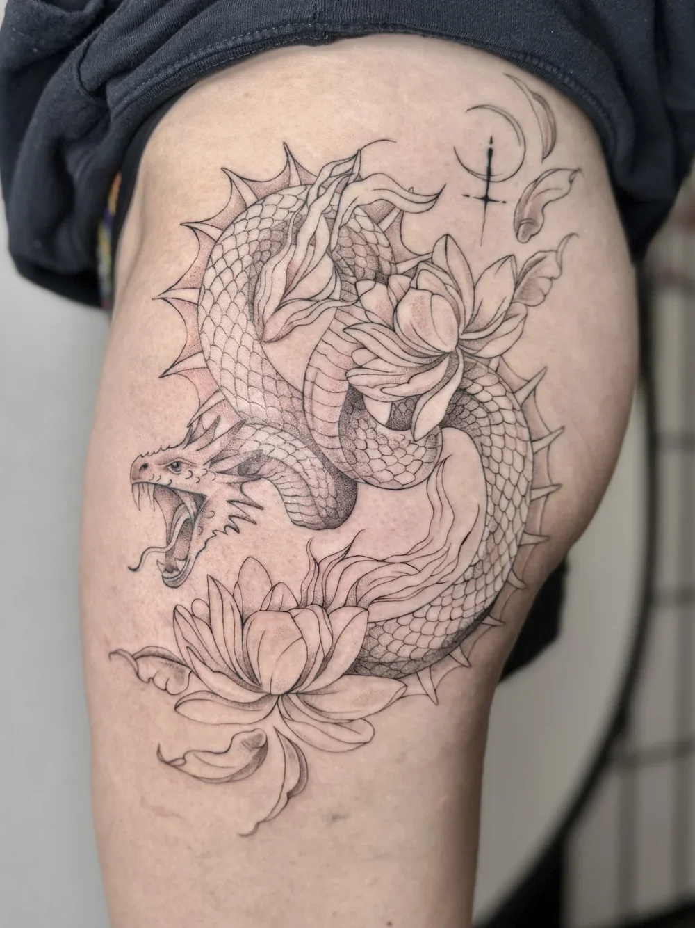 Black and gray tattoo of a coiled snake with an open mouth, detailed scales, and fangs, surrounded by large blooming lotus flowers, a moon, a moon phase symbol, and a small celestial object.