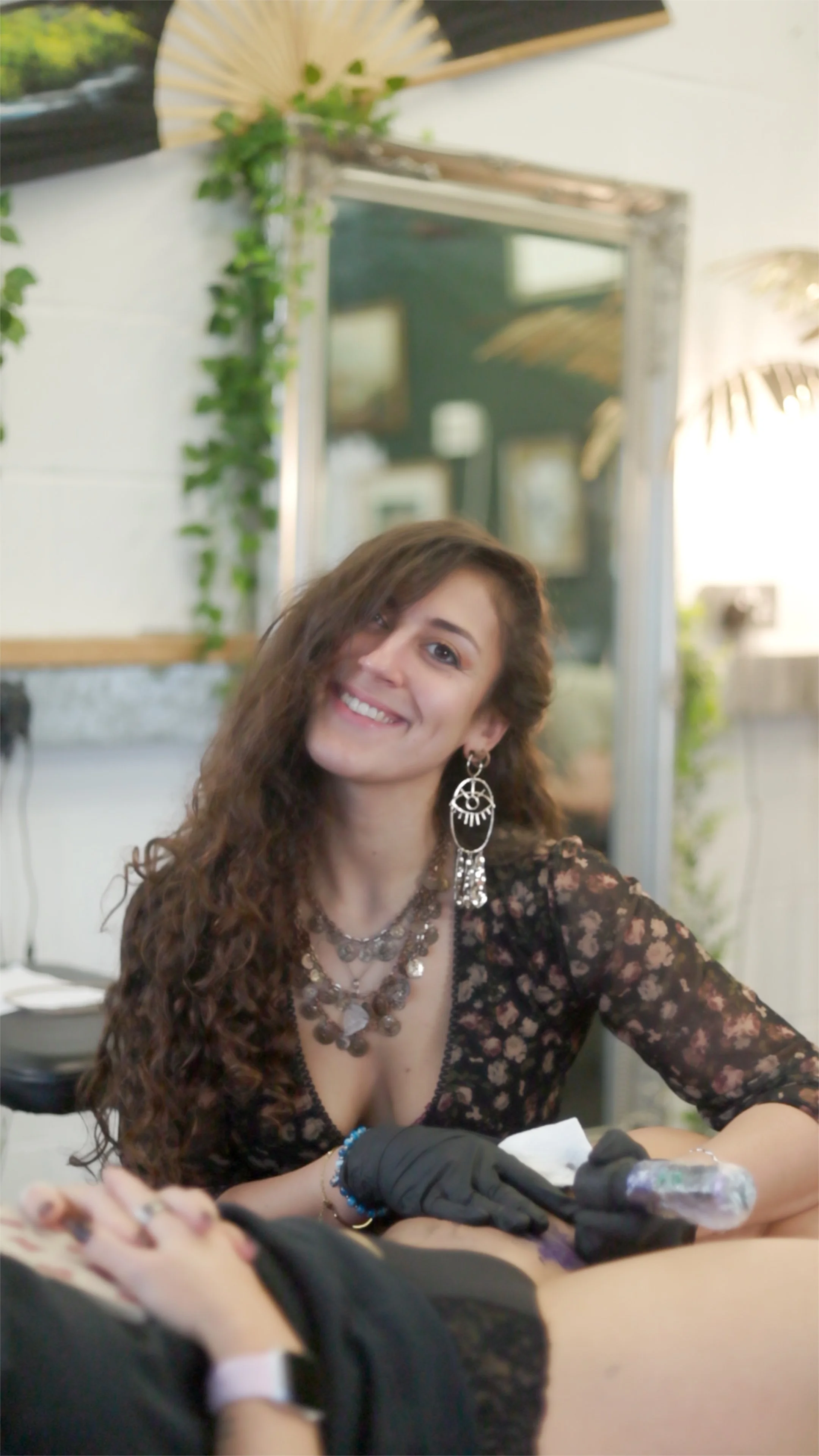 A woman with long, curly brown hair and large earrings, smiling while tattooing someone.