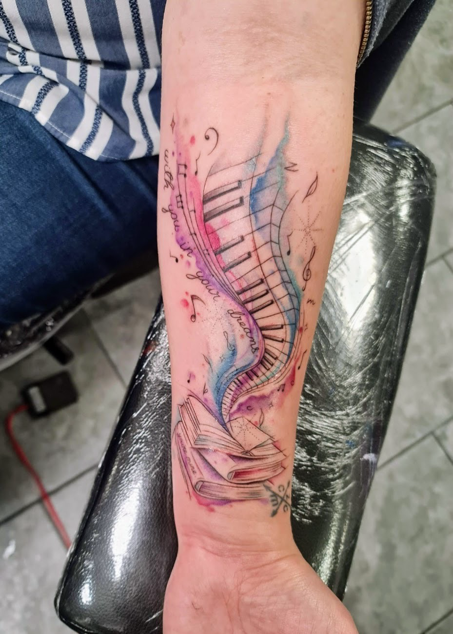 Colorful tattoo of a piano keyboard, musical notes, and an open book on a person's forearm.