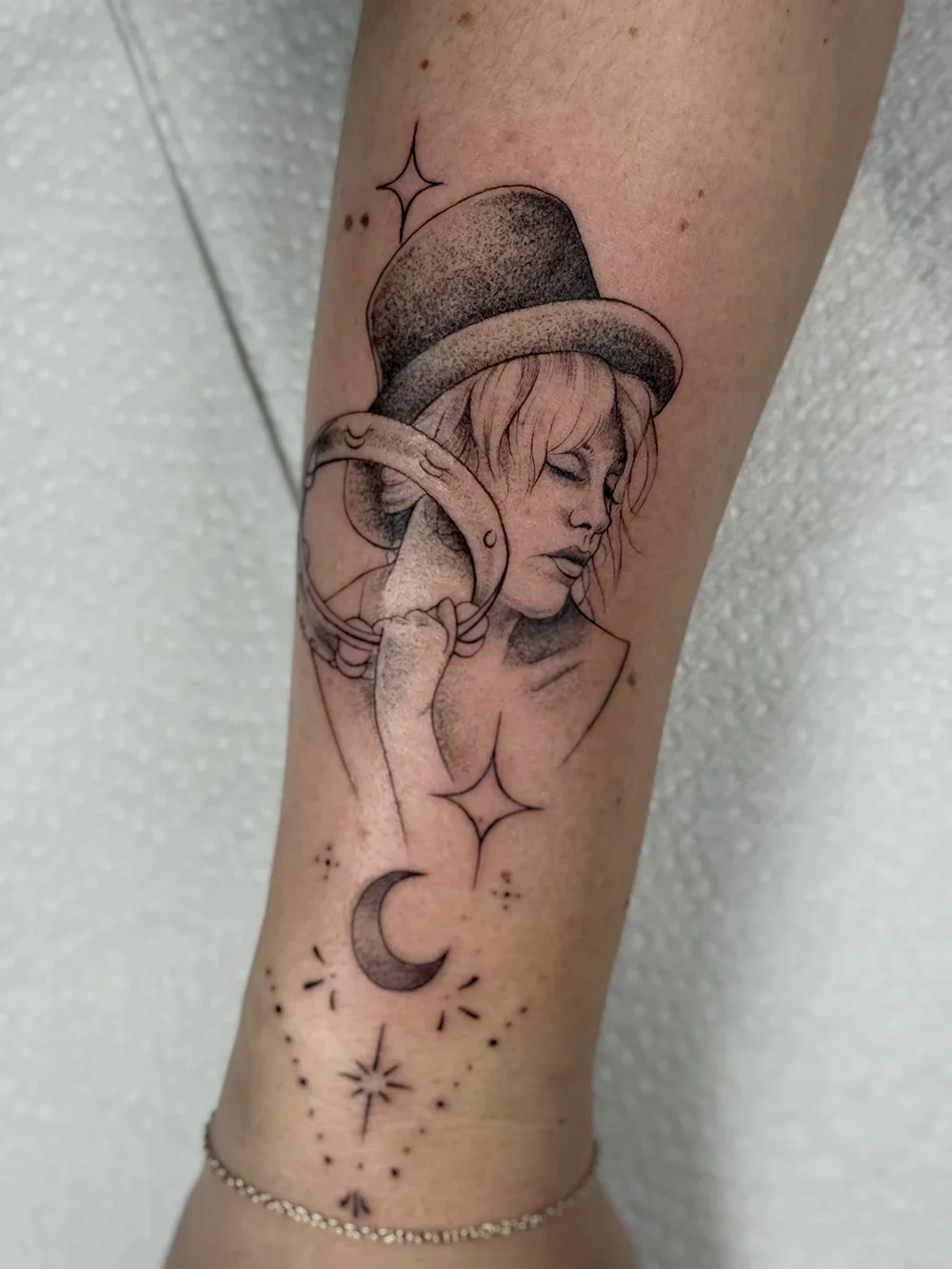 Tattoo of a woman with closed eyes wearing a hat, holding a ring, with stars, a crescent moon, and small decorative elements below.