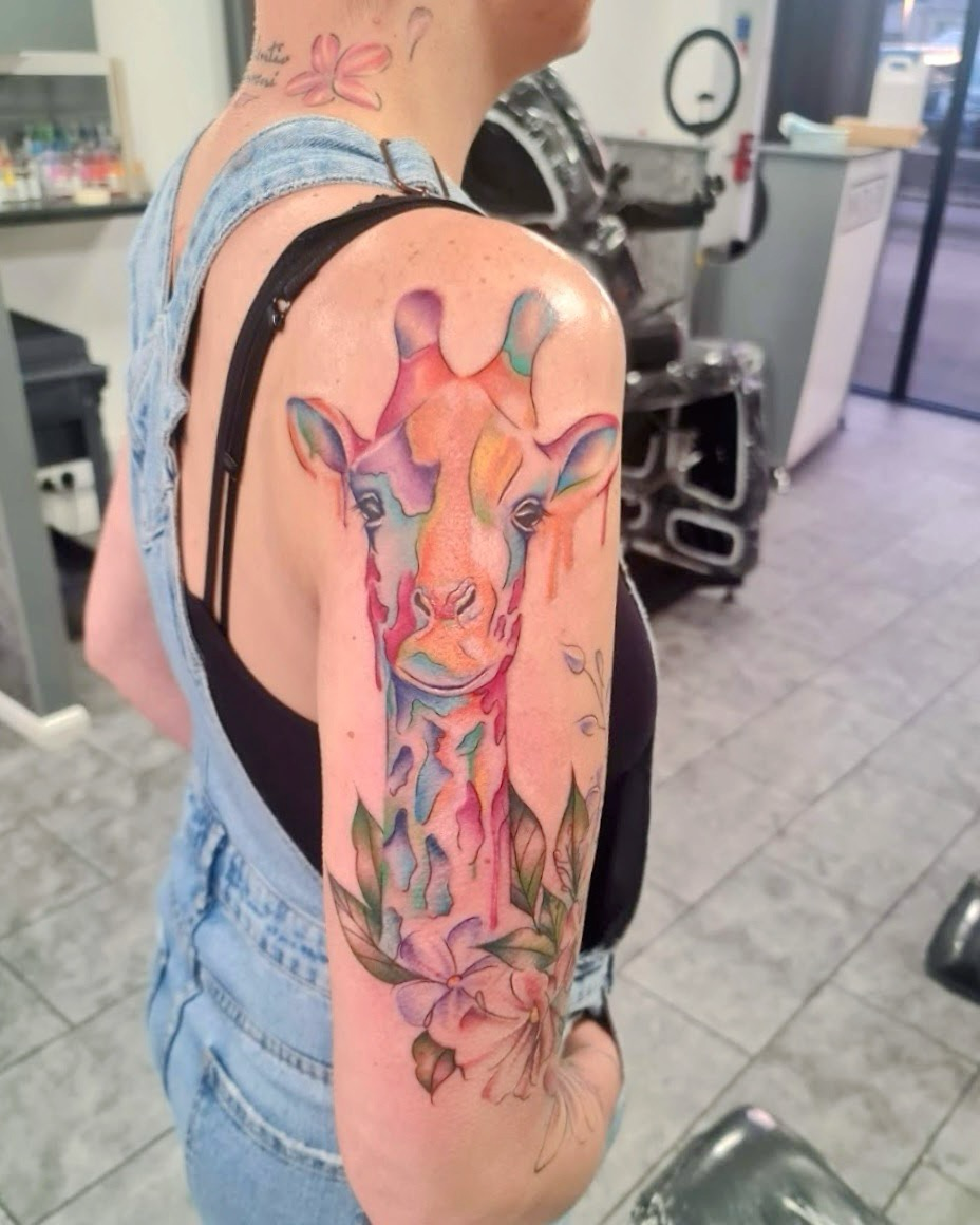 Colorful watercolor-style tattoo of a giraffe's face and neck, with flowers and leaves, on a woman's upper arm.
