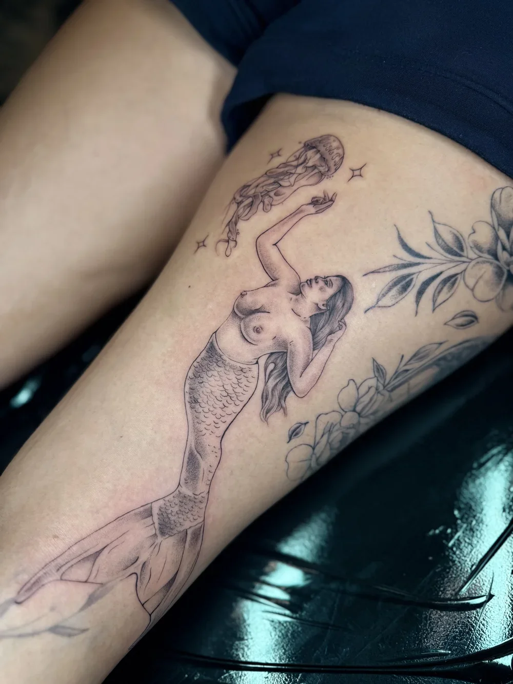 Tattoo of a mermaid with long hair, holding her hair with both hands, surrounded by flowers, with a floating figure of a woman with closed eyes and flowing hair above her.