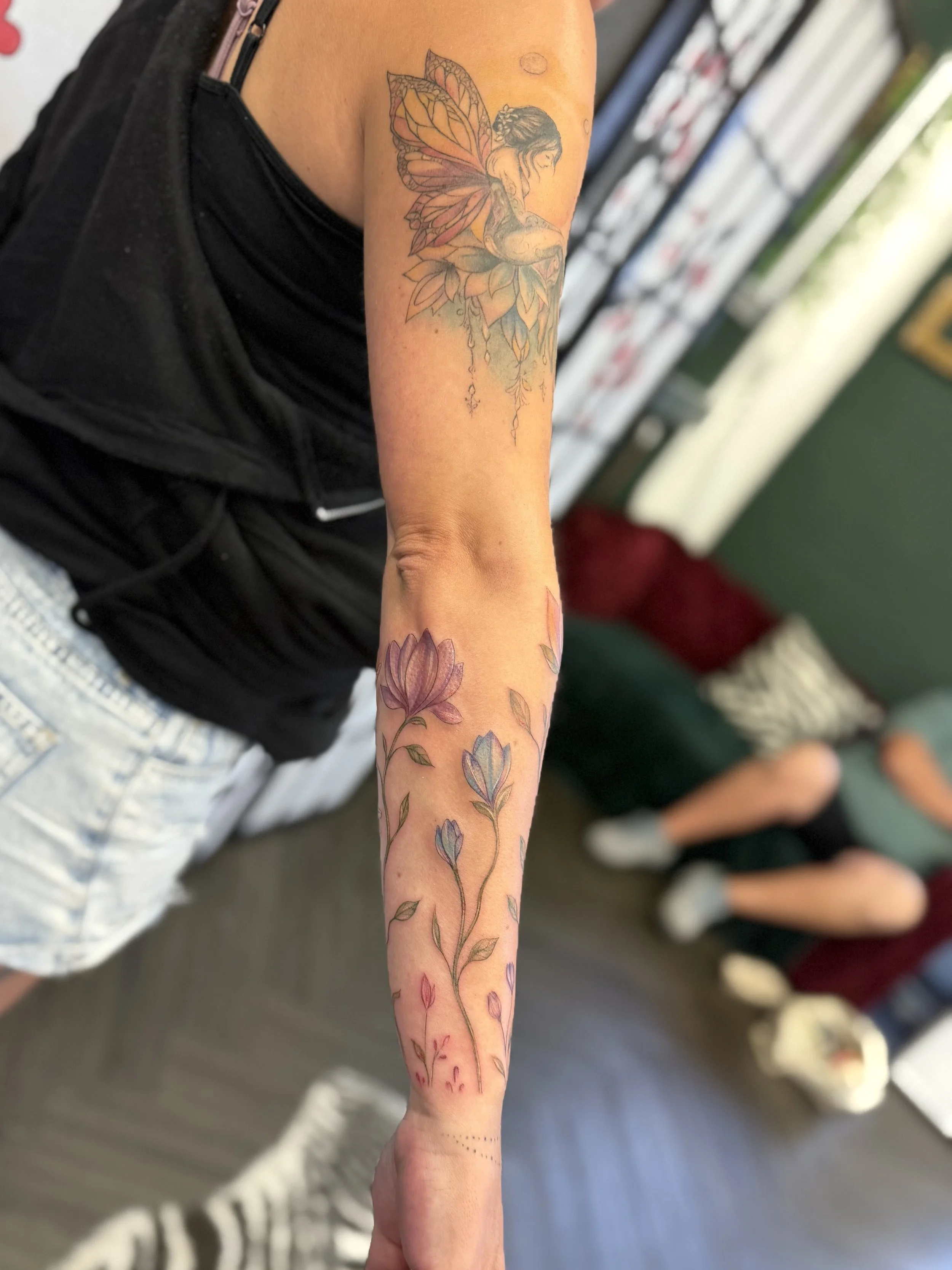 Tattoo of a fairy with butterfly wings sitting on a large flower with flowing hanging leaves on the upper arm, and a flowering plant with purple and pink flowers running down the forearm.