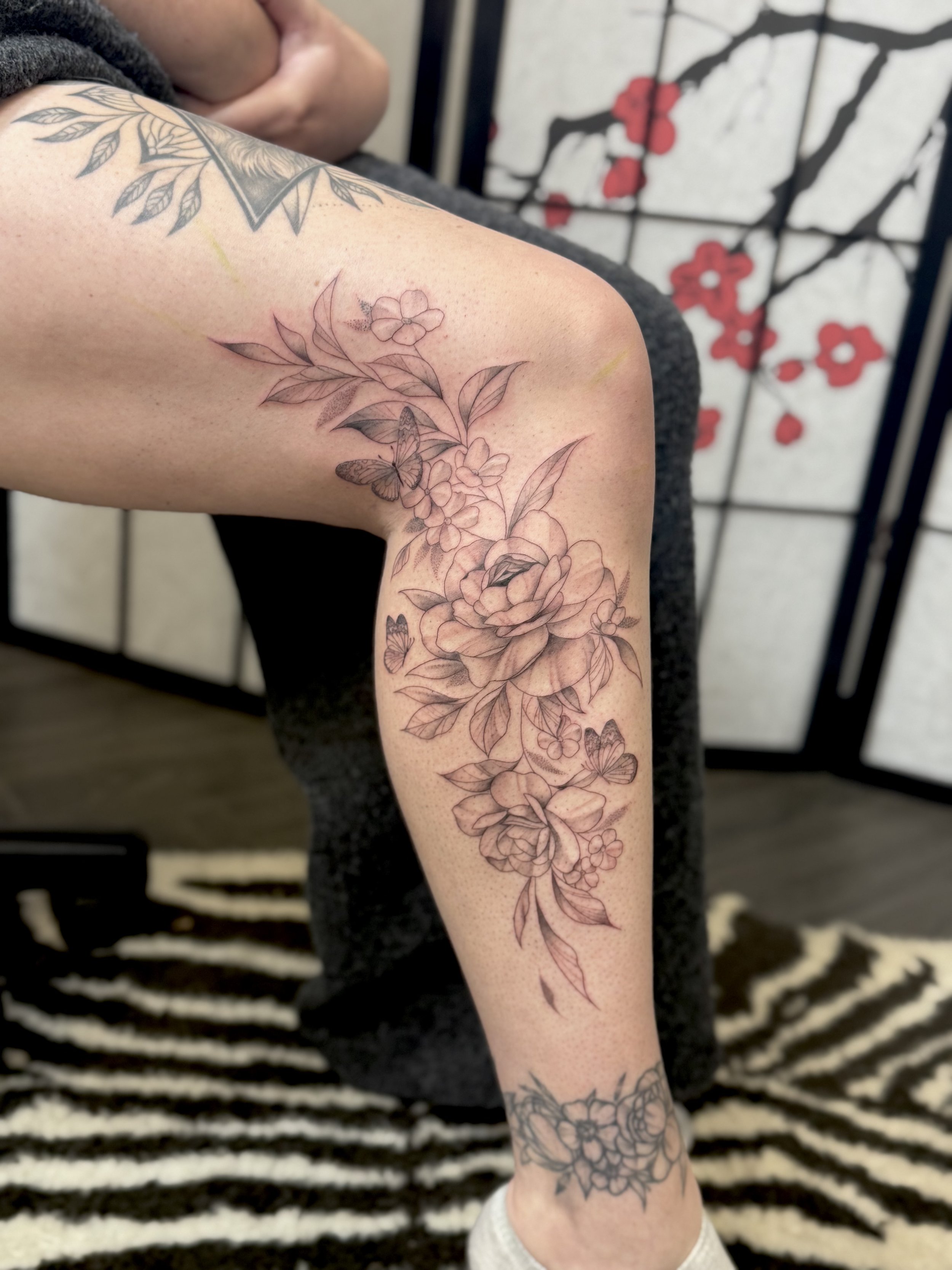 A person's leg featuring a large floral tattoo with blossoms, leaves, and butterflies, in progress, in a studio with decorative screen behind.