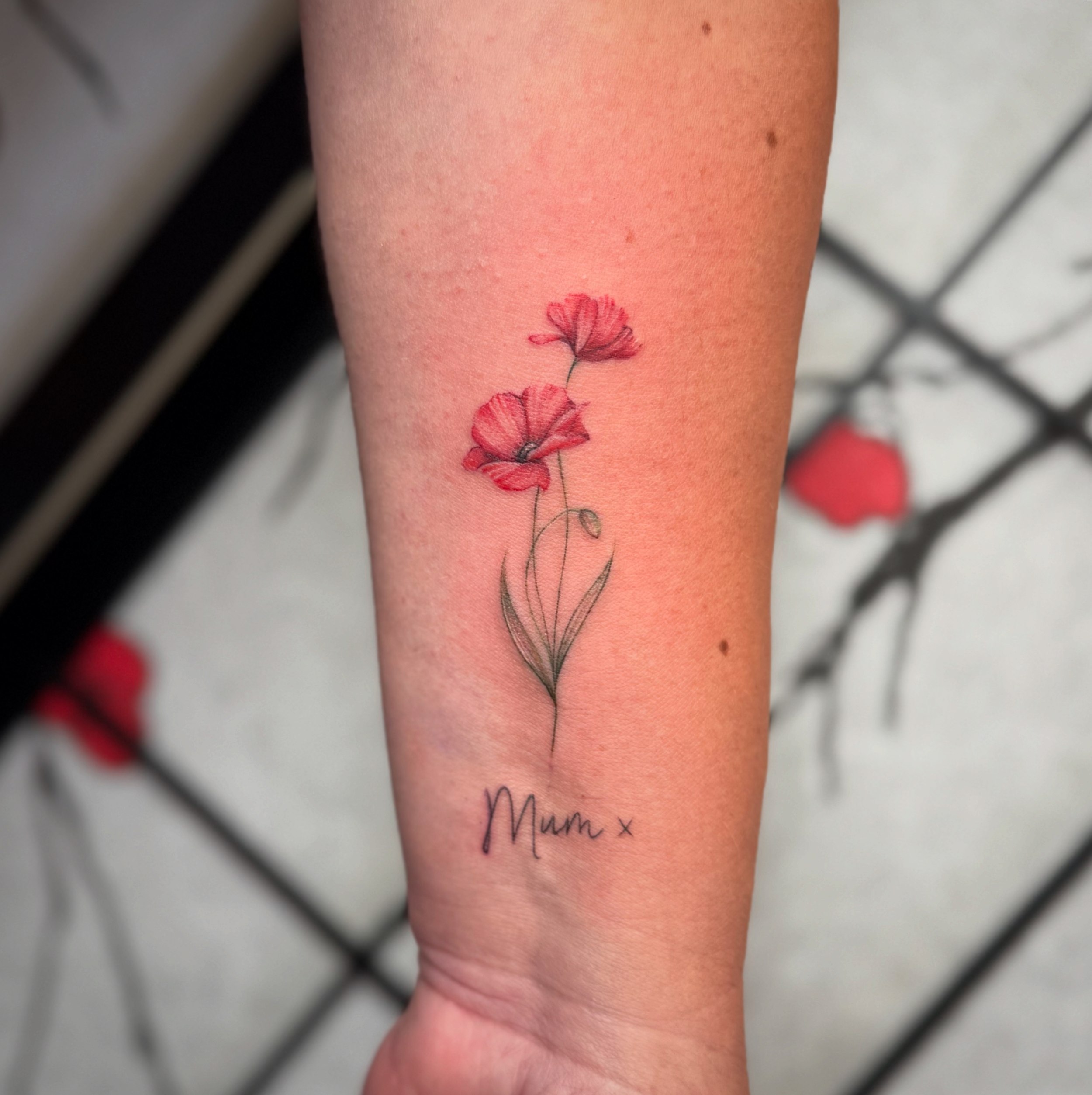 A tattoo of two pink poppy flowers with green stems and leaves, and the word 'Mum' with a small 'x' underneath, on the person's forearm.