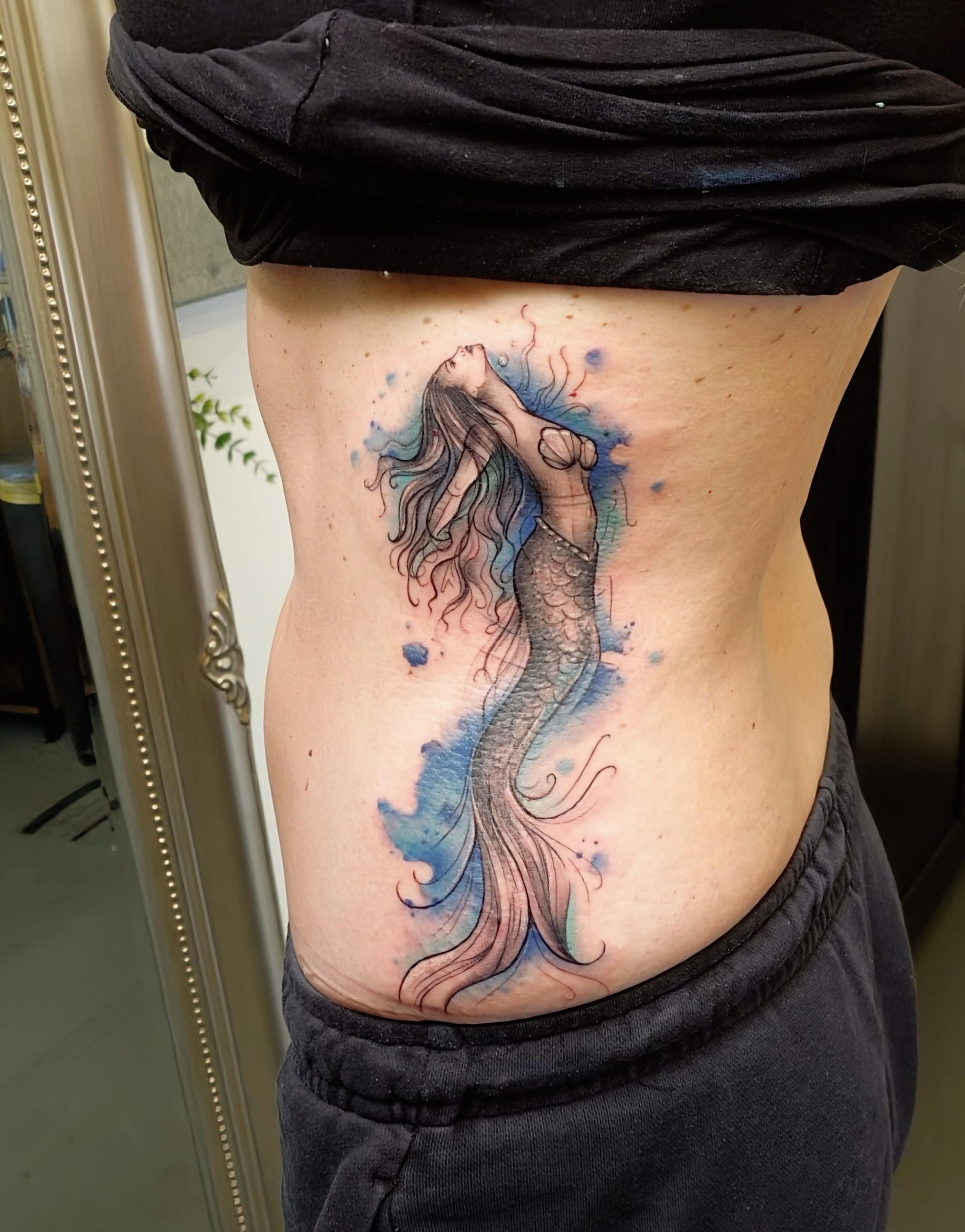 Tattoo of a mermaid on a person's side, with long flowing hair and a fish tail, surrounded by blue watercolor splashes.