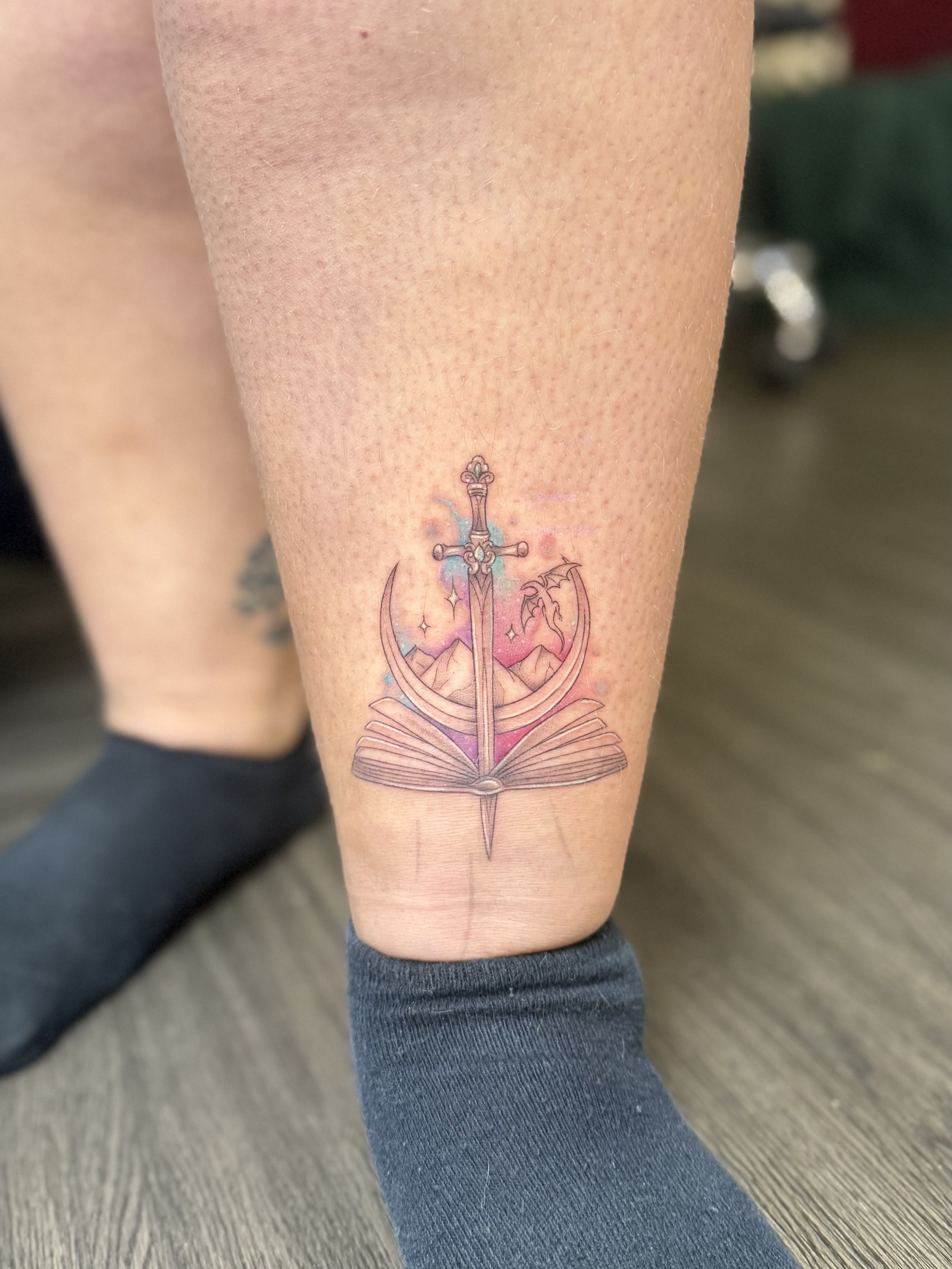 Tattoo of a sword, a crescent moon, an open book, mountains, and a dragon on a person's leg.