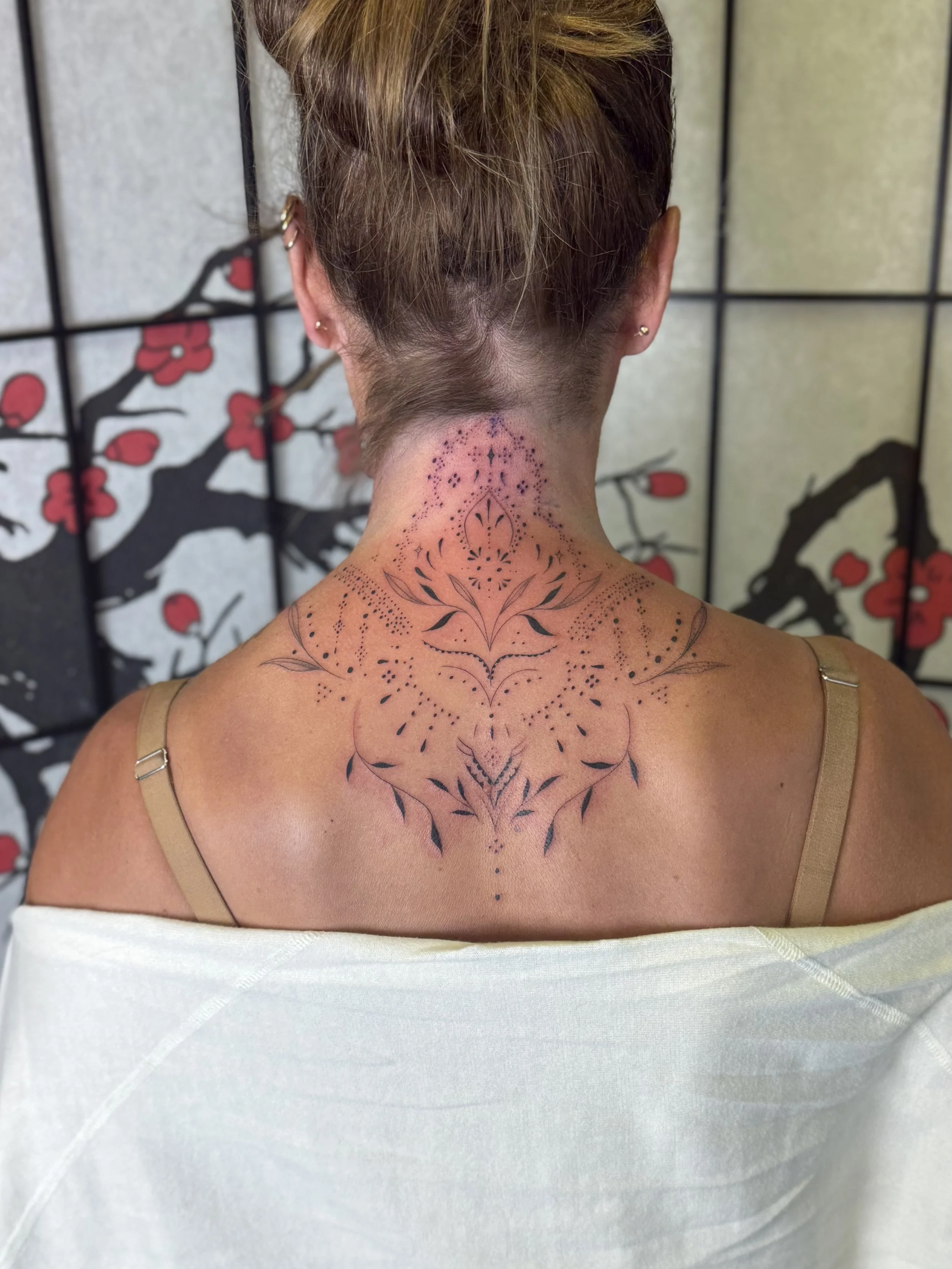 Back of a woman with a tattoo on her neck and upper back, featuring black ink tribal and floral designs, with some red and purple ink areas.