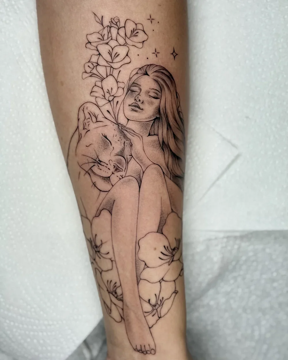 Tattoo of a woman with long hair, her eyes closed, embracing a lioness, surrounded by flowers and stars.