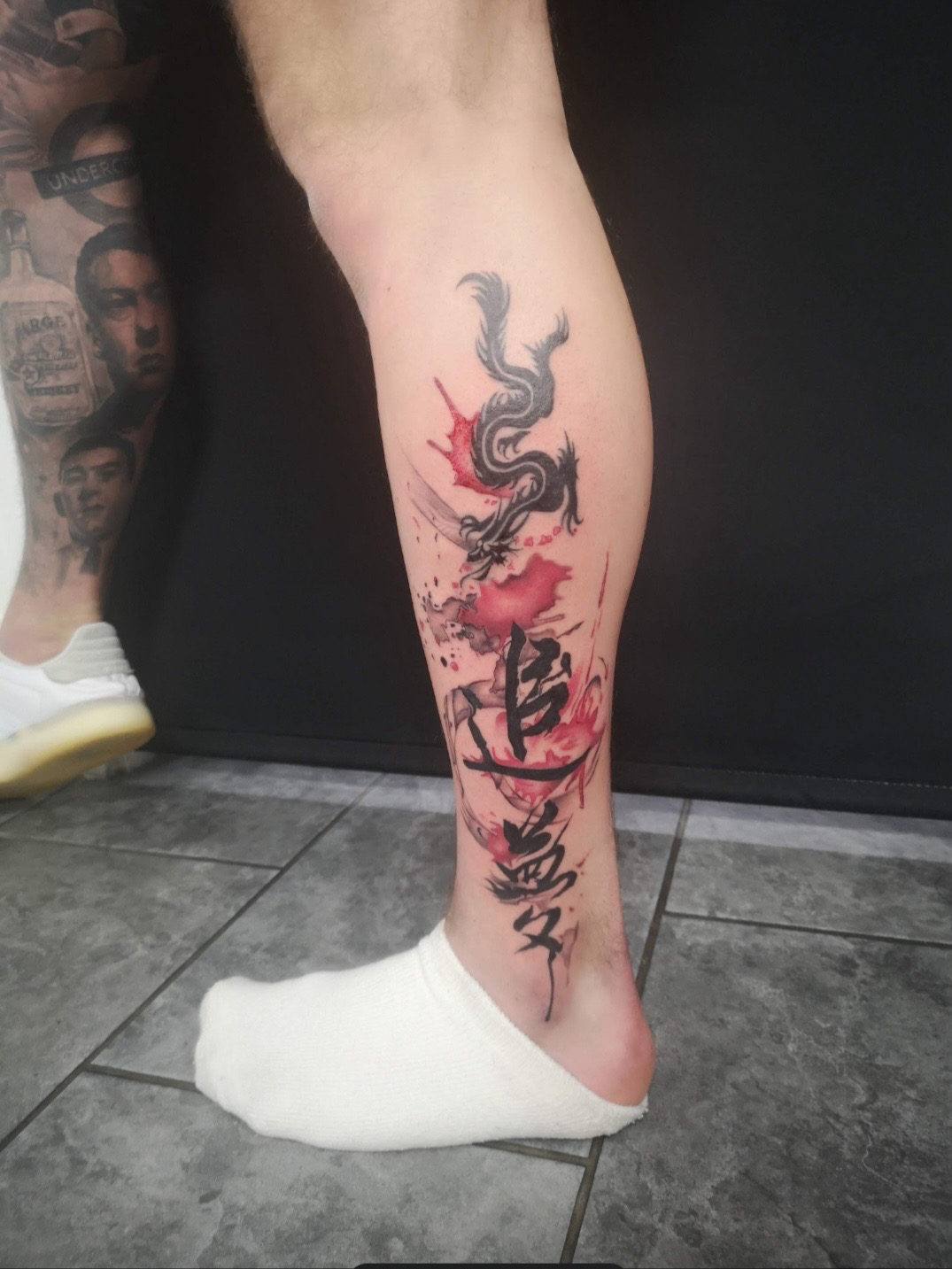 Tattoo of a black dragon with red accents on a person's lower leg, with ink splashes and Japanese characters.