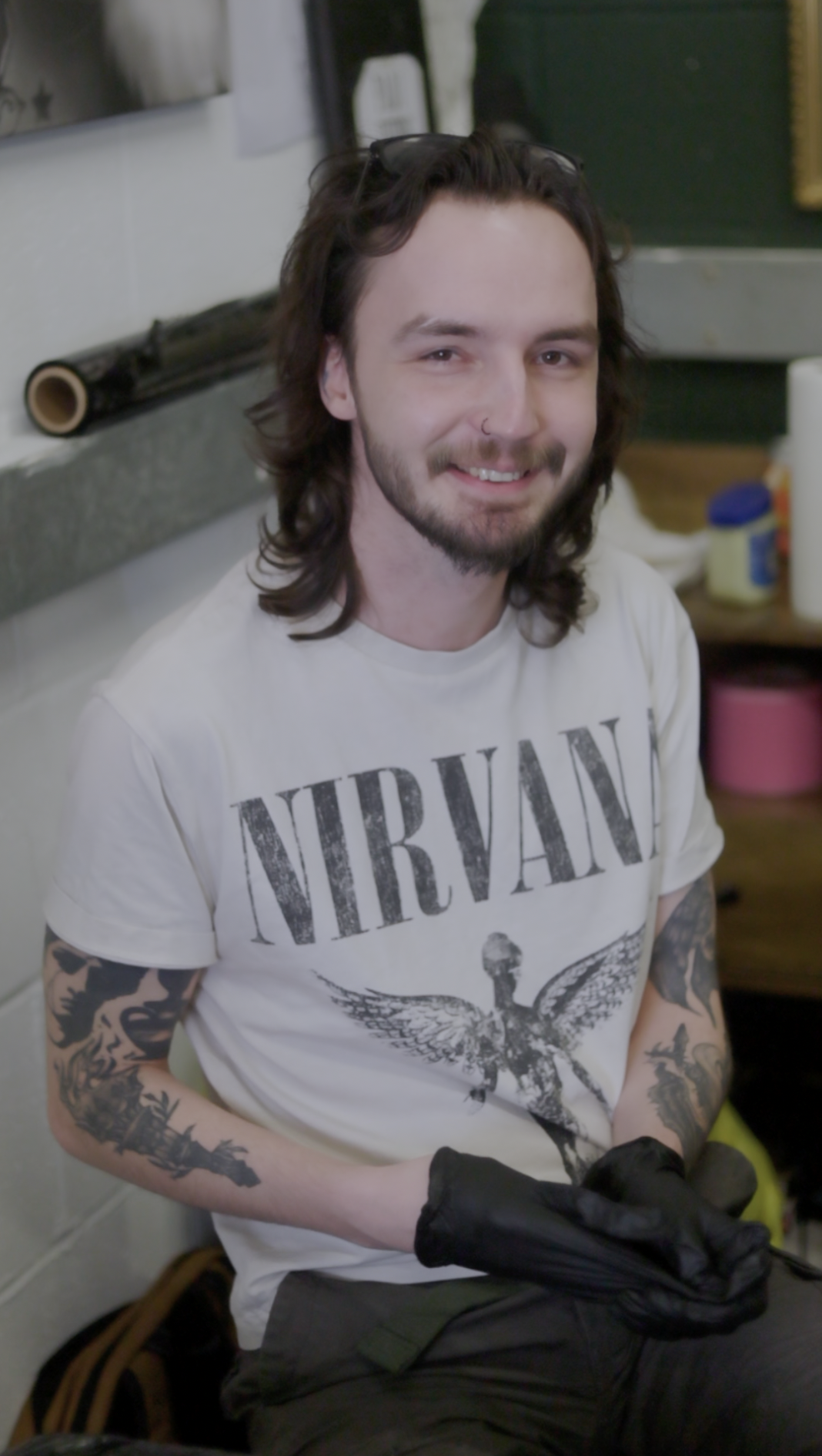 A man with long brown hair, a beard, and tattoos on his arms, wearing a Nirvana T-shirt, sitting indoors and smiling at the camera.