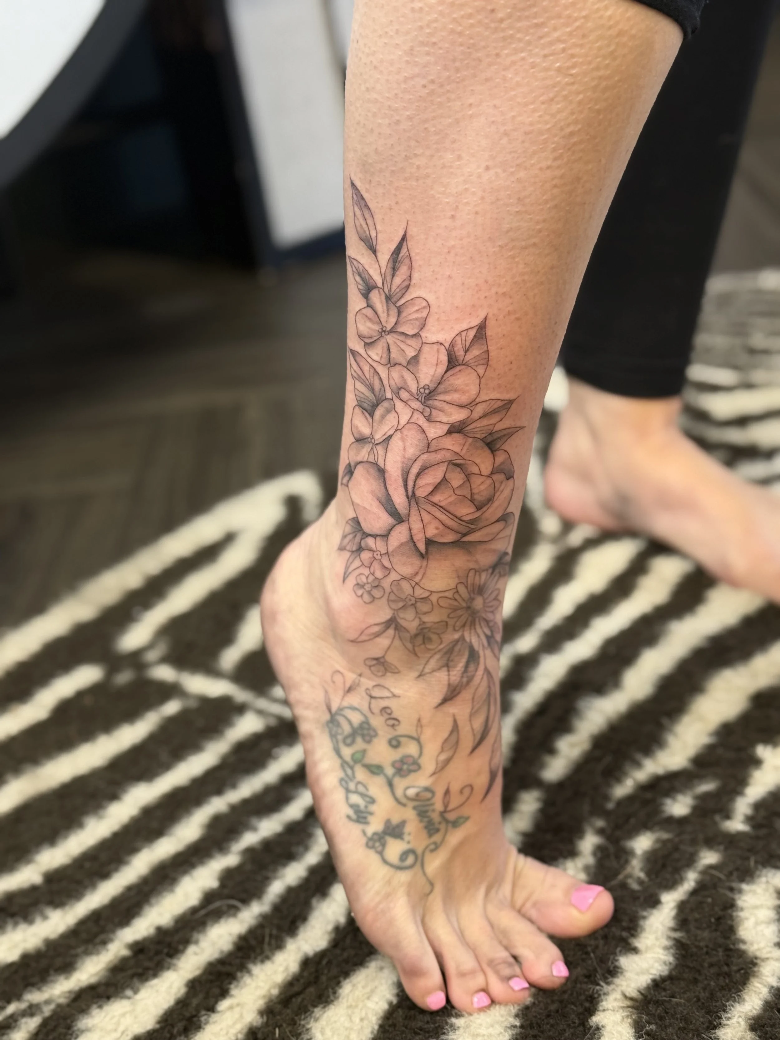 A person's leg with a floral tattoo extending from the ankle to the mid-calf, featuring a large rose and other smaller flowers and leaves. There is also script writing on the foot.