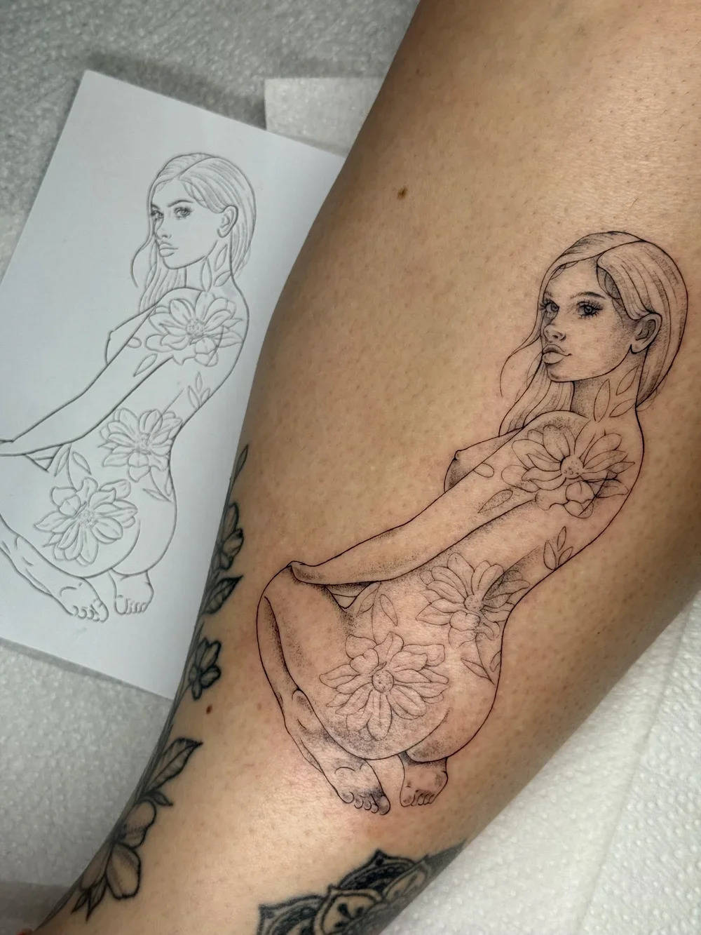 Tattoo artist working on a floral woman tattoo design on a person's leg, with a reference sketch nearby.