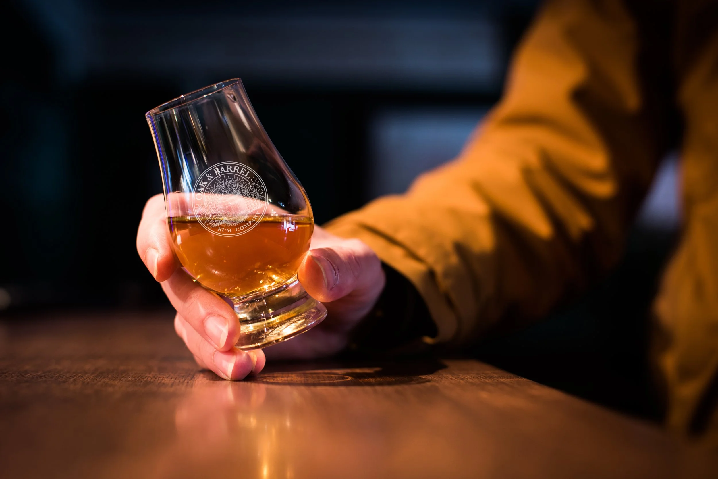 A hand holding a glass of amber-colored liquor with a logo that reads 'Oak & Barrel Rum Company.'