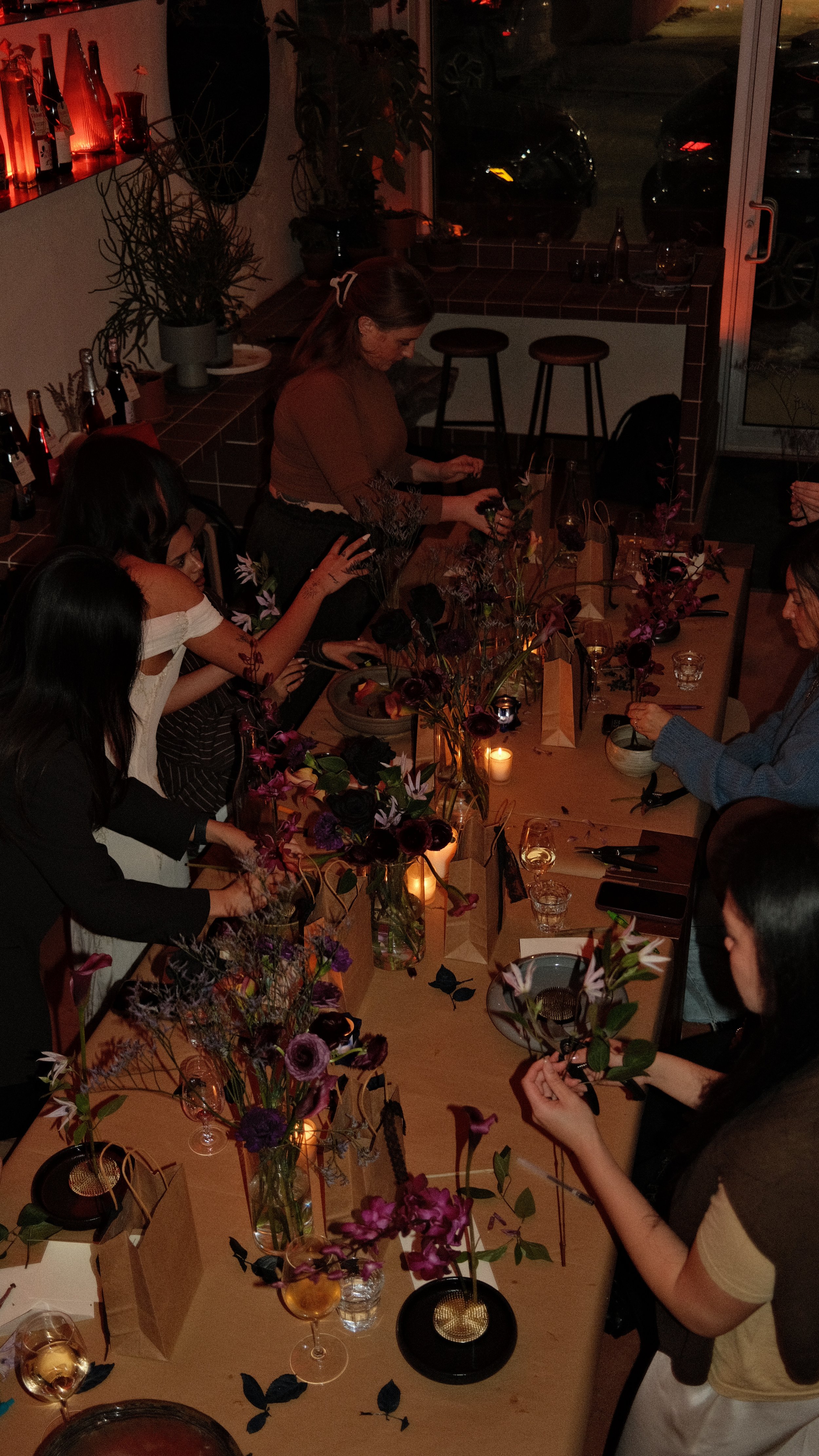 People gathered around a long banquet table decorating floral arrangements in a dimly lit room with candles and wine.