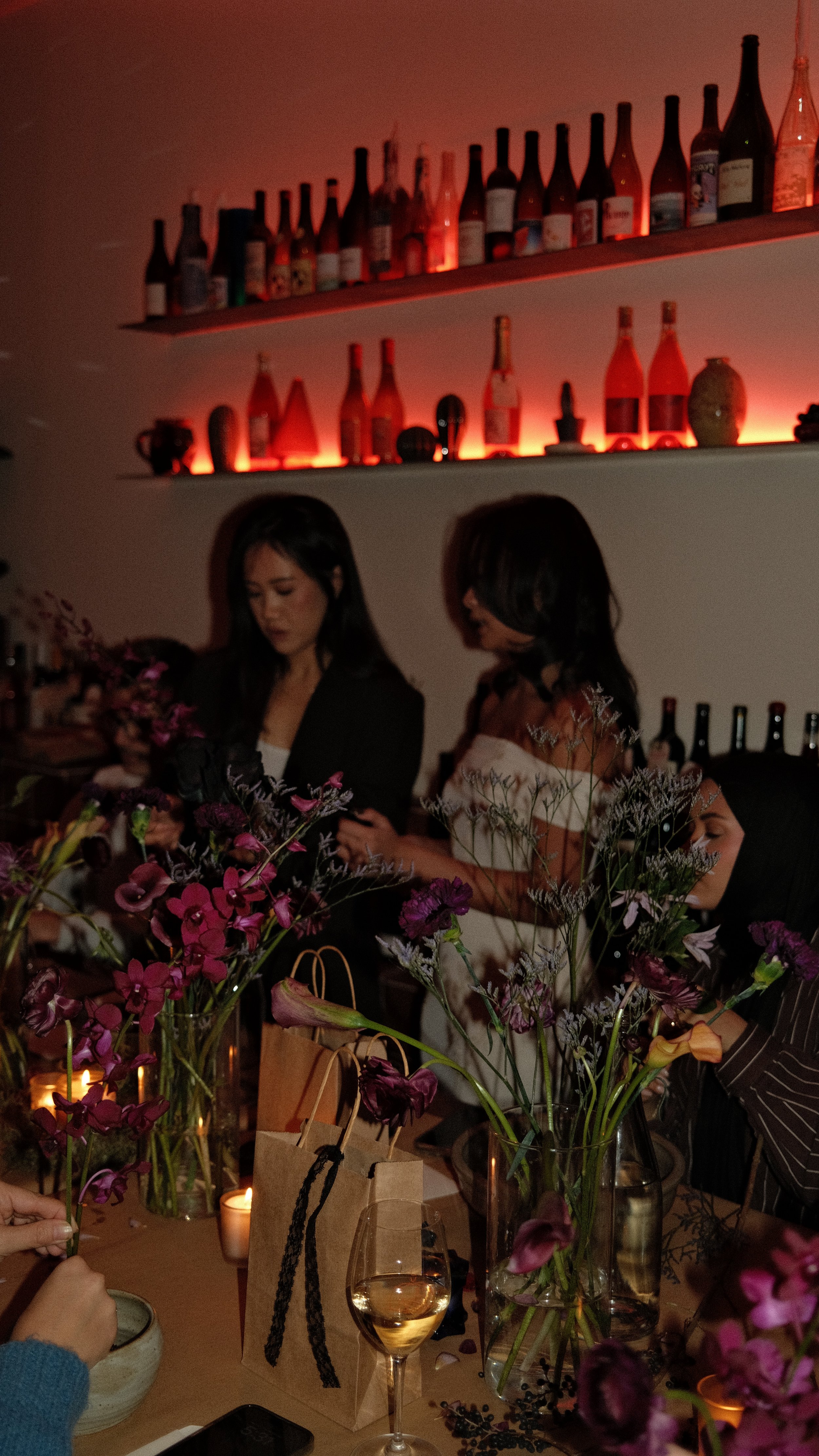 People gathered around a table with pink and purple flowers, wine glasses, and candles, in a dimly lit room with bottles on shelves in the background.