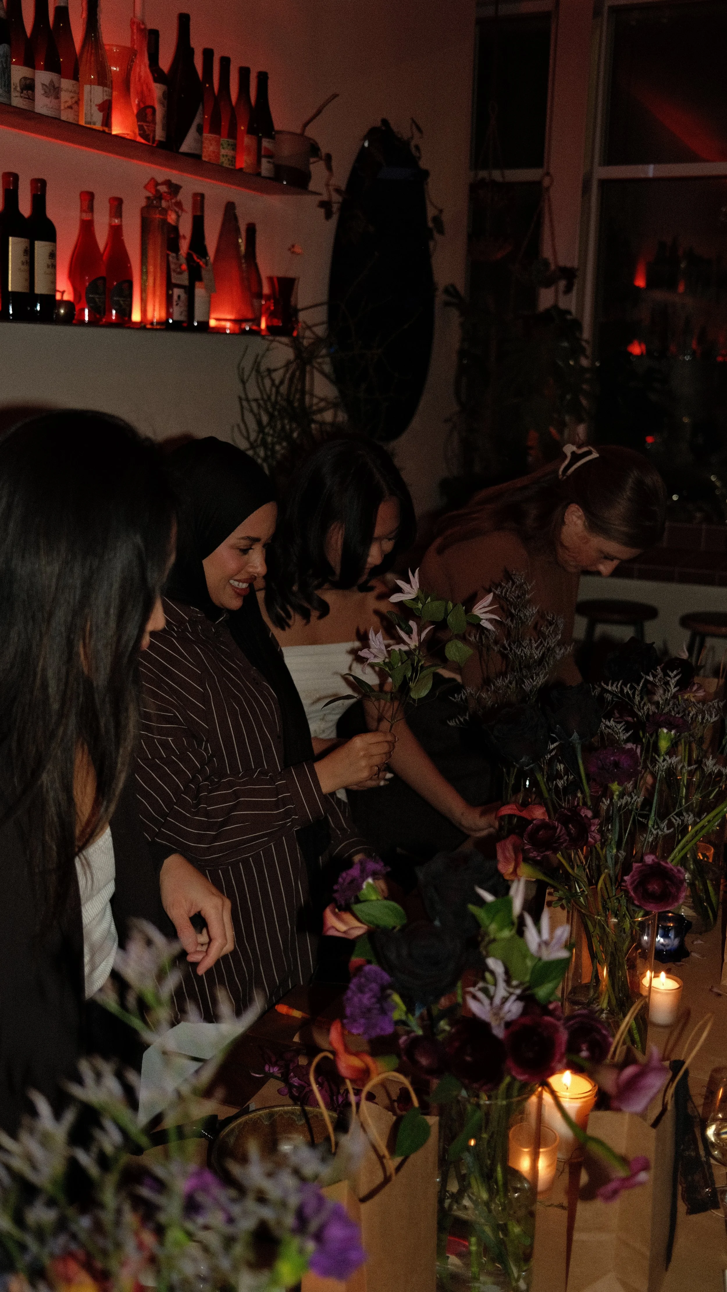 Women arranging flowers on a table with lit candles, in a dimly lit room with bottles on shelves in the background.