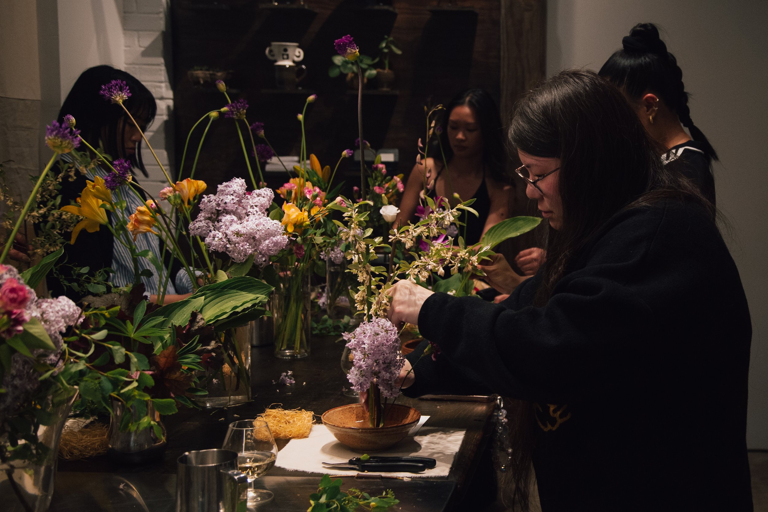 Group of women arranging and creating floral arrangements with various colorful flowers in a dimly lit room.