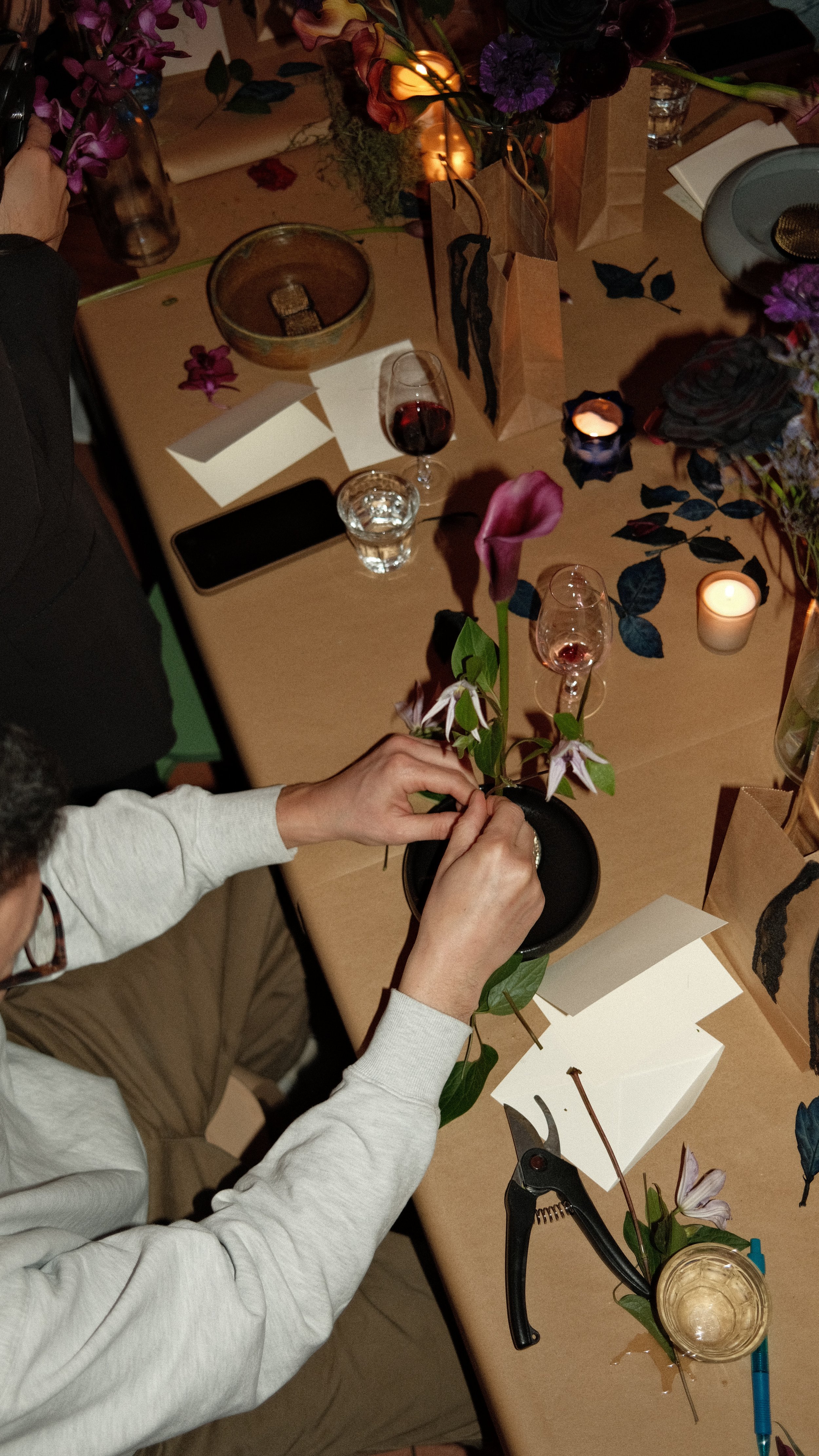 Person arranging a flower in a black pot on a decorated table with candles, glasses, and gift bags.