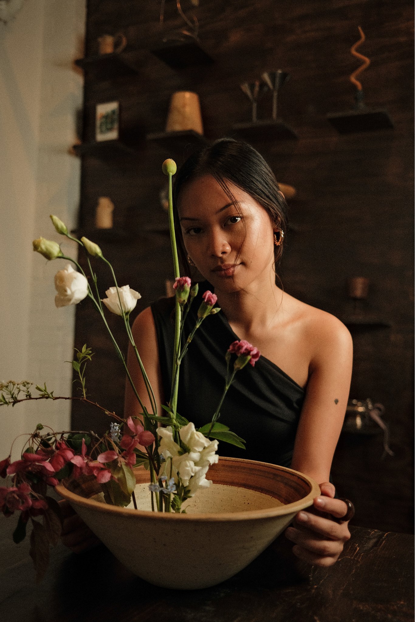 A woman with black hair and gold earrings arranging flowers in a large bowl on a dark wooden table in a cozy, dimly lit room.