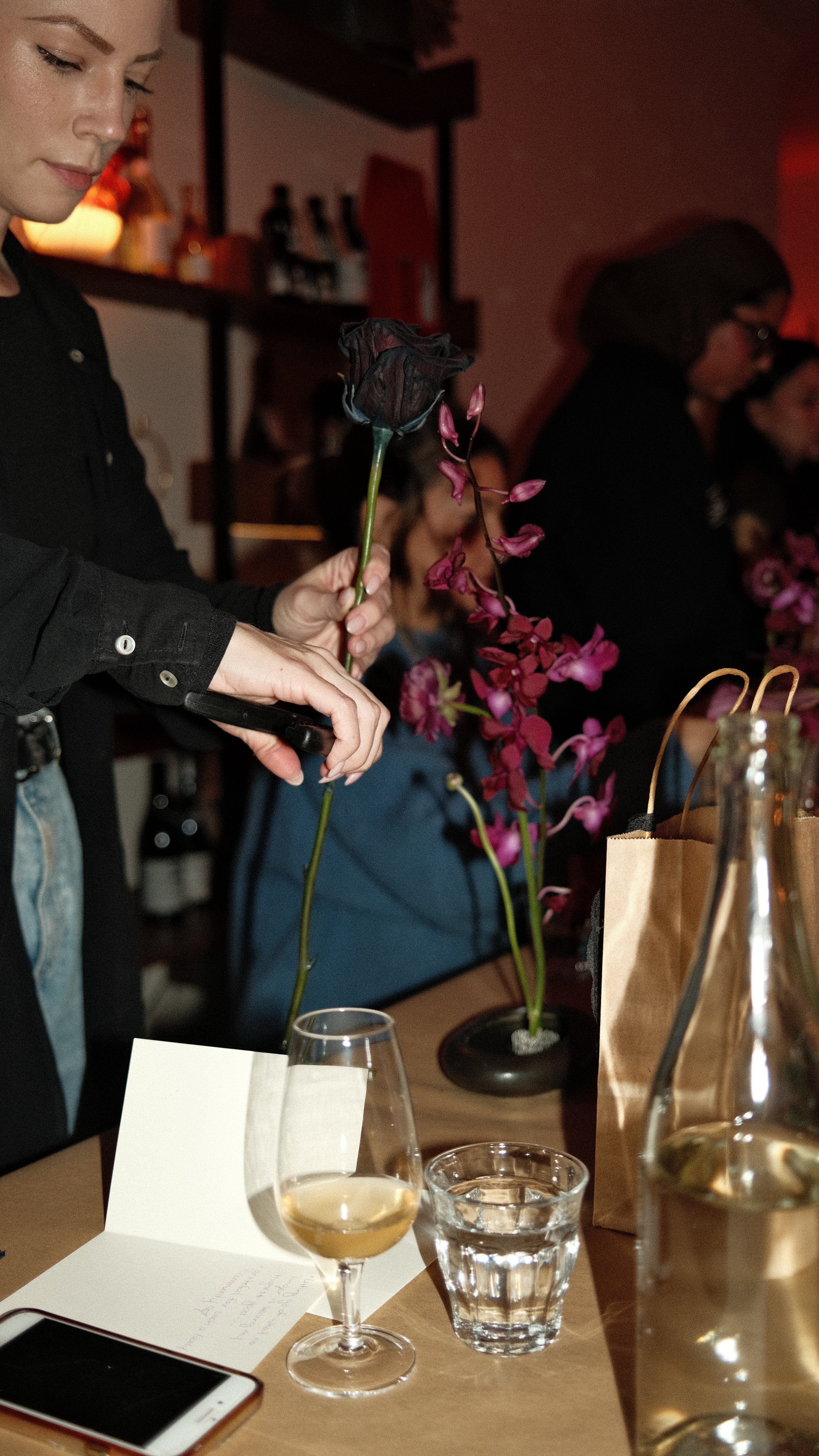 Person holding a black rose and pink orchids at a table with wine, water, and a paper bag in a dimly lit setting with people in the background.