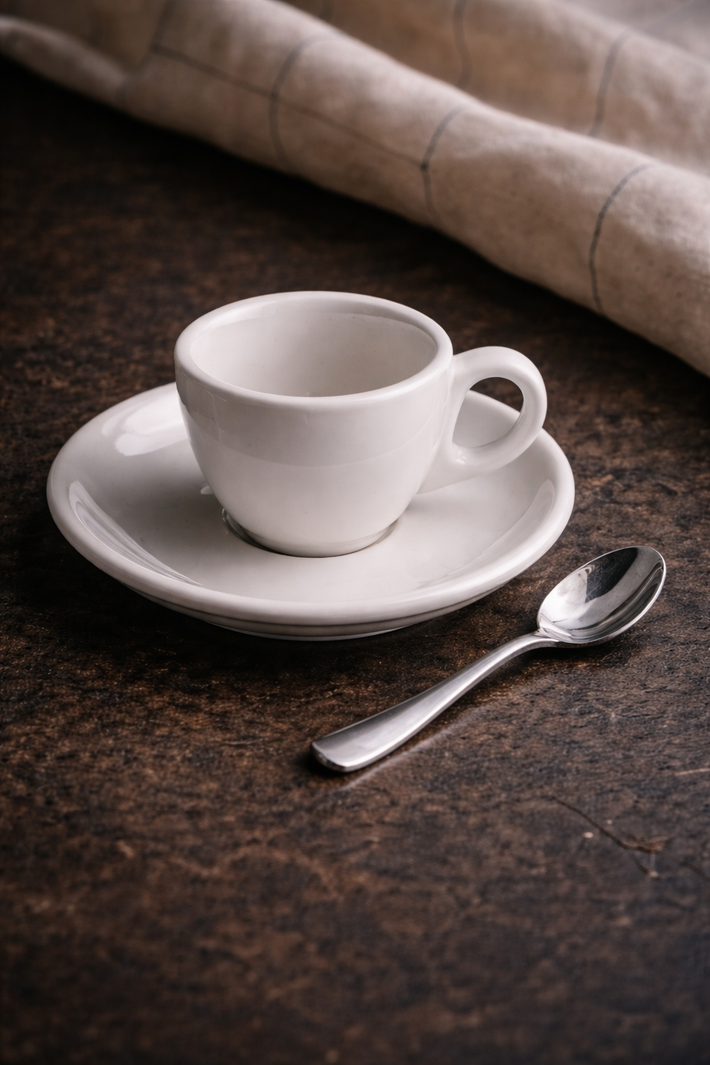 Weiße Kaffeetasse auf einem weißen Unterteller mit Löffel auf einem dunklen Holz Tisch, im Hintergrund beige Stoff.